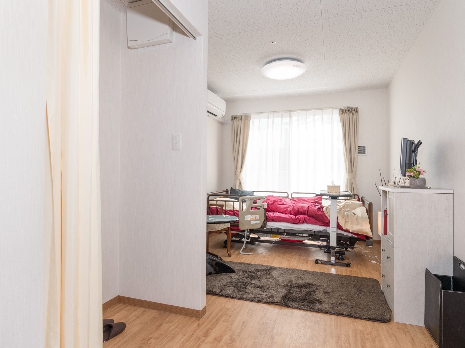 左手前に、カーテンで仕切られるトイレがある。室内にはエアコンや介護ベッド、チェストなどが置かれている。