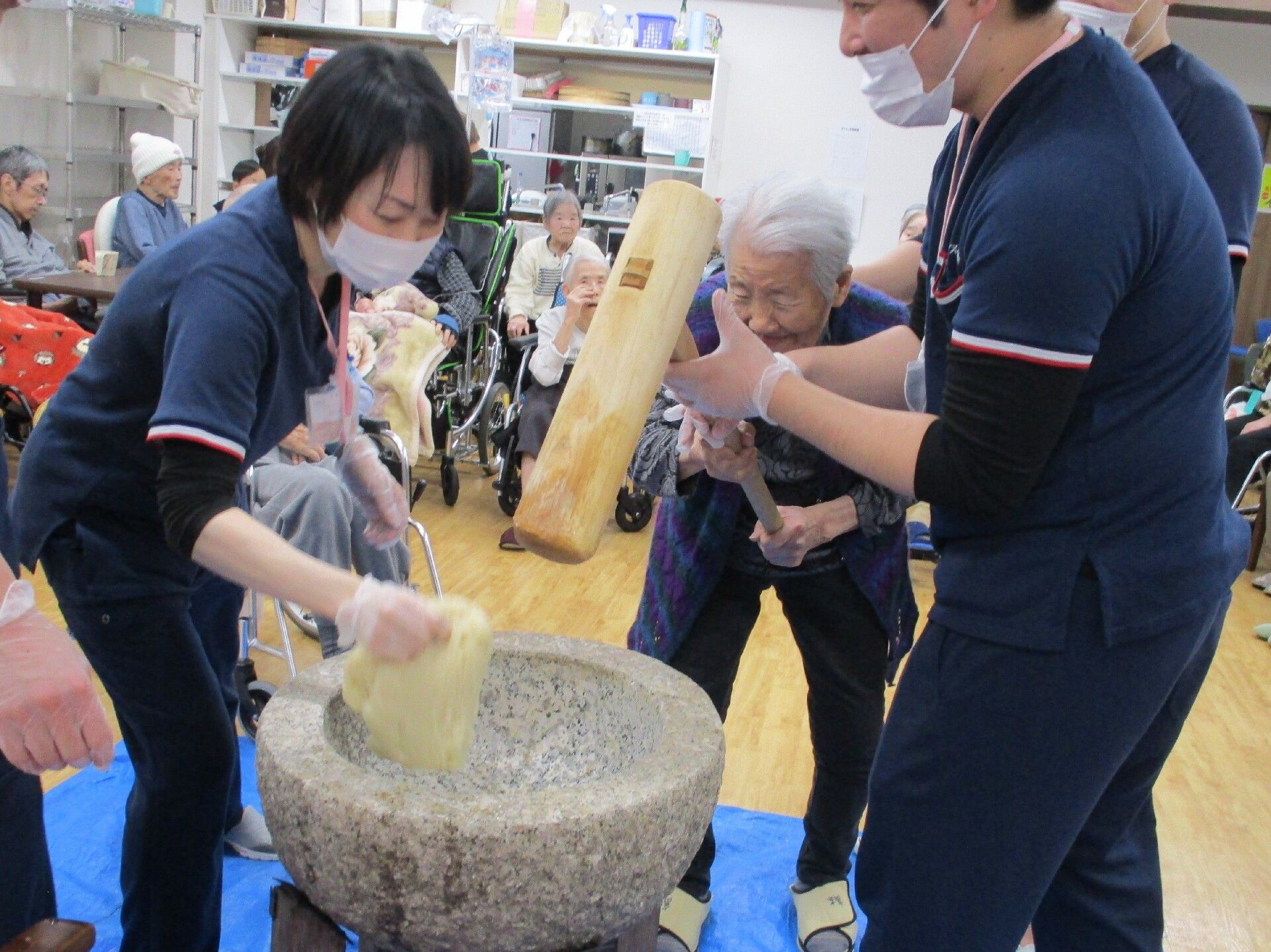 スタッフと入居者が杵とうすを使用し、一緒に餅つきを楽しんでいる様子。室内で行っている。