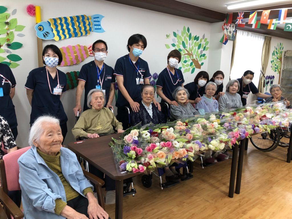 多数の花束が並べられたテーブルに着席している入居者様と後ろに立つスタッフ