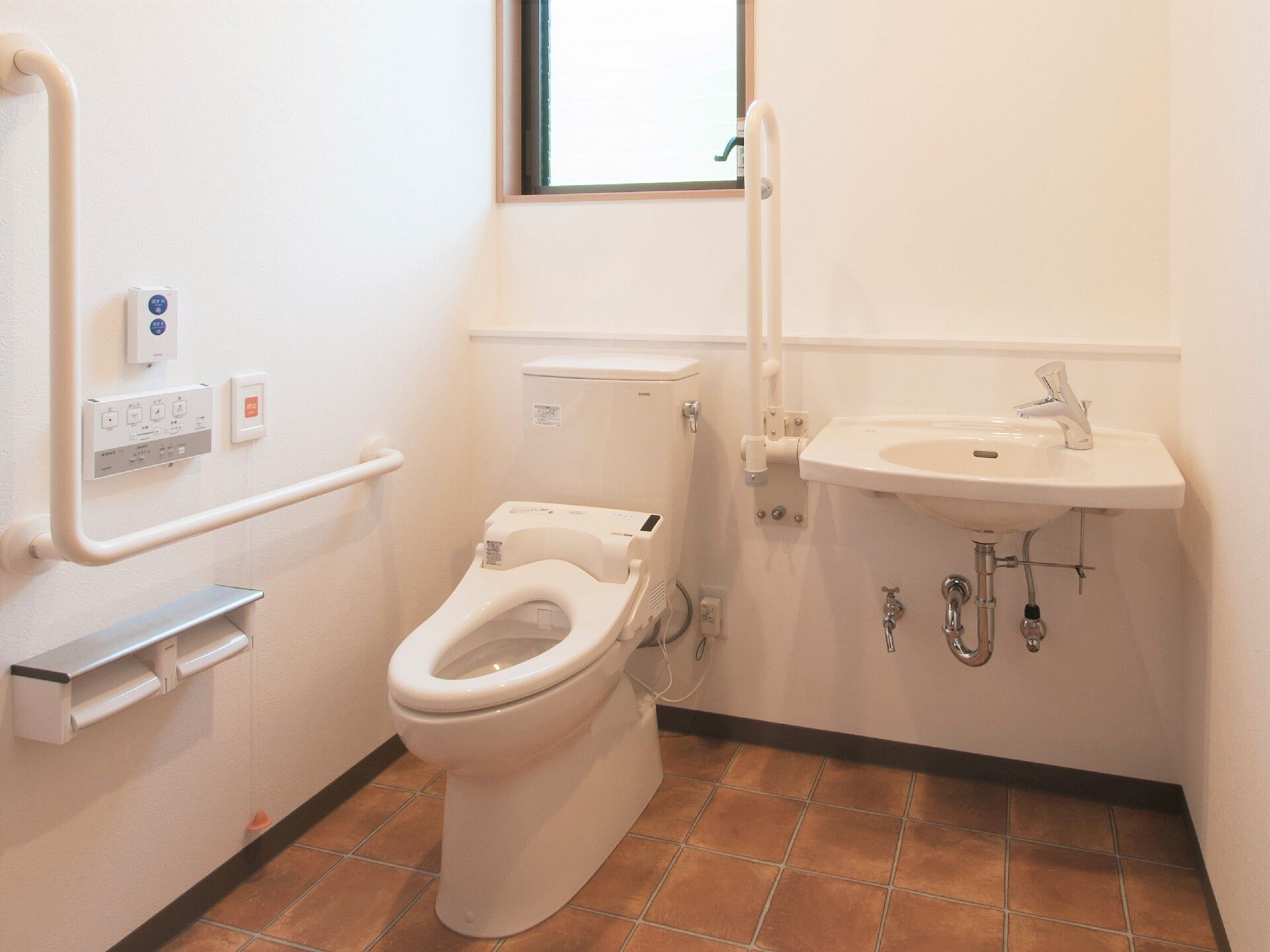 広いトイレにはふたなしの便座が付いたトイレと手洗い場があり、壁にはエル字型の手すりや呼び出しボタンなどが付いている。