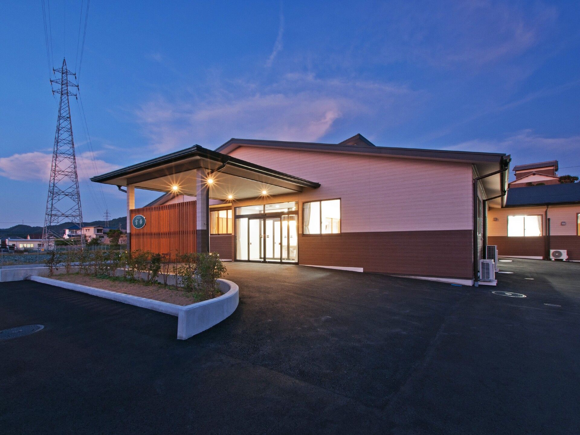 三角屋根が付いた平屋建ての建物。玄関には大きな車寄せが付き、背の低い木が植えられたスペースもあり、夕暮れ時でライトアップされている。