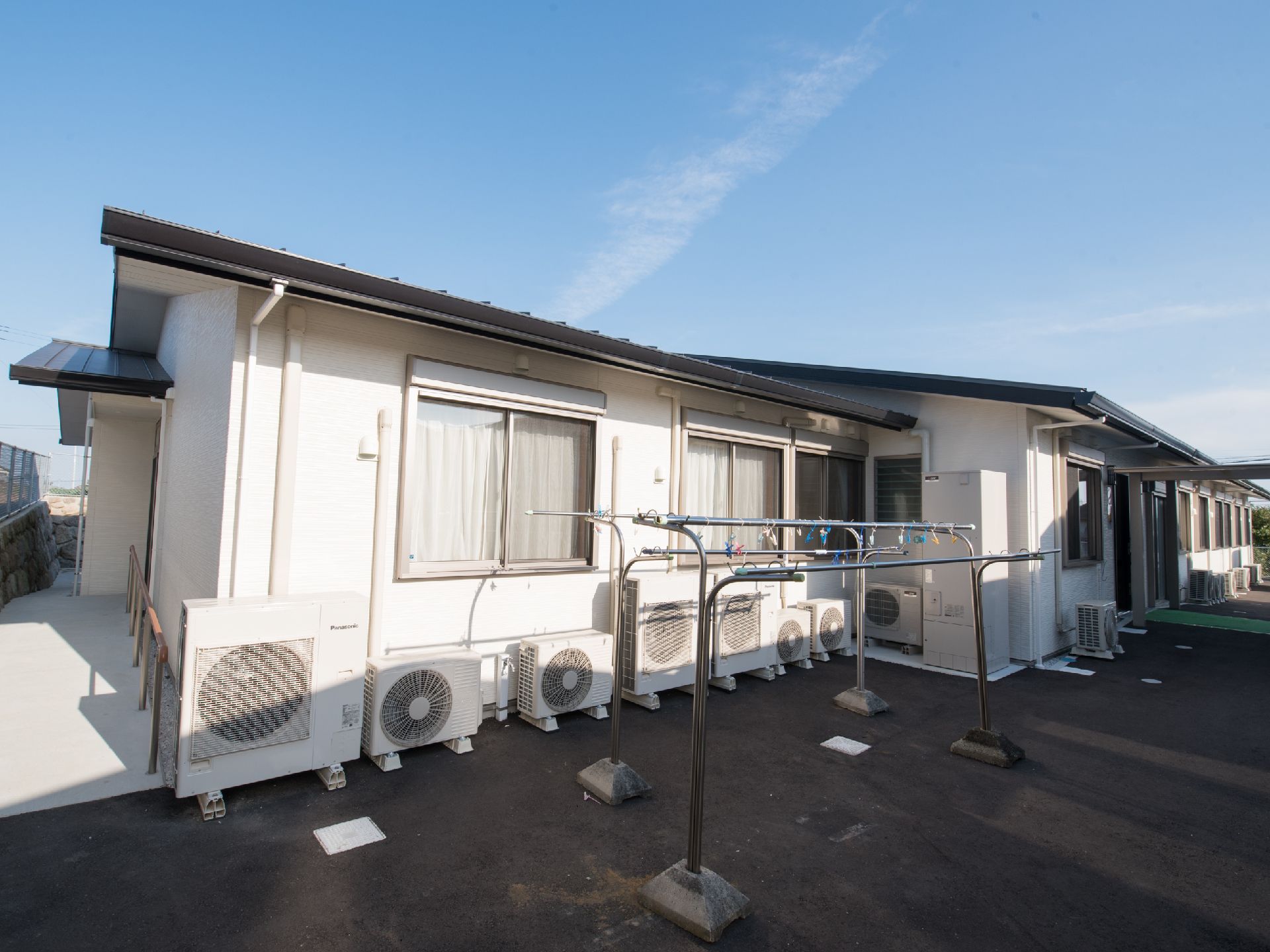 施設の建物は平屋である。白色の壁には居室の窓が並び、地面にはエアコンの室外機が置かれている。洗濯物干しの設備がある。