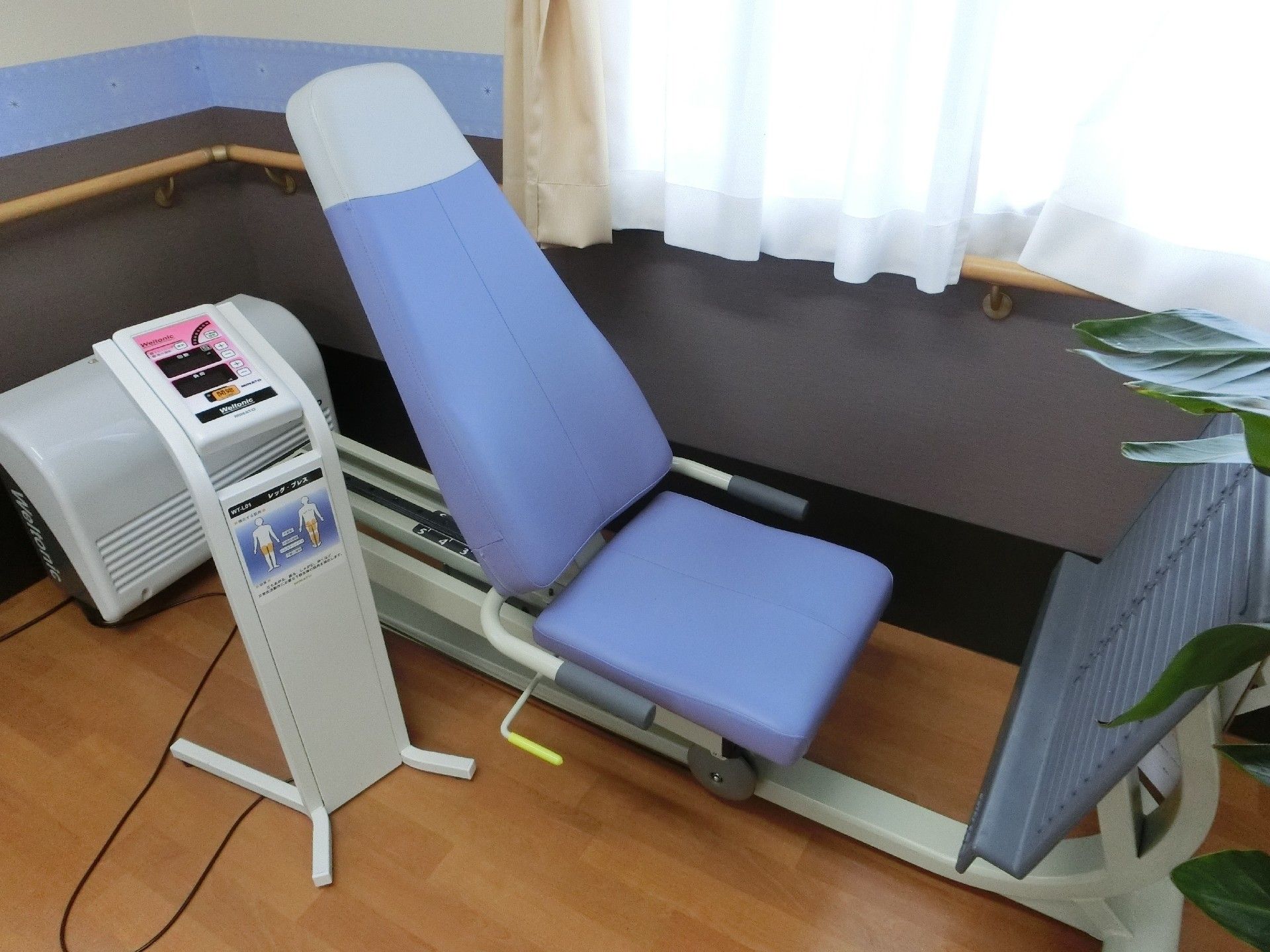 部屋の隅に青色のレッグプレスが置かれている。リハビリスペースの窓には、レースと白色のカーテンが取り付けられている。