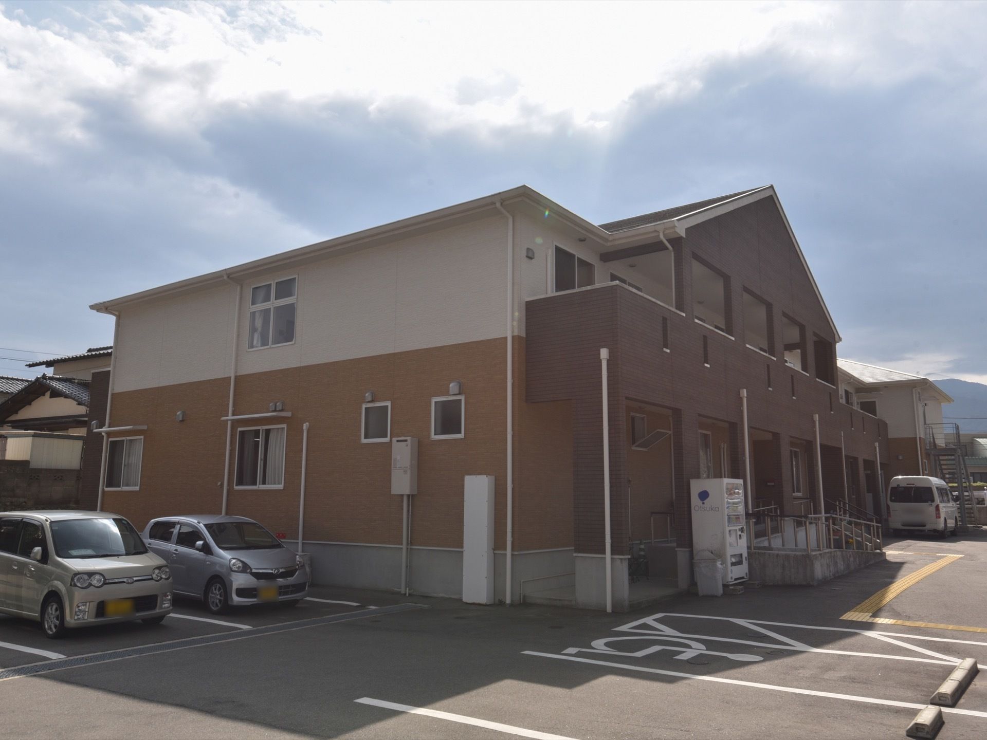 地上２階建ての施設を、斜め方向から写したものである。左横には駐車スペースがあり、軽自動車が２台置いてある。