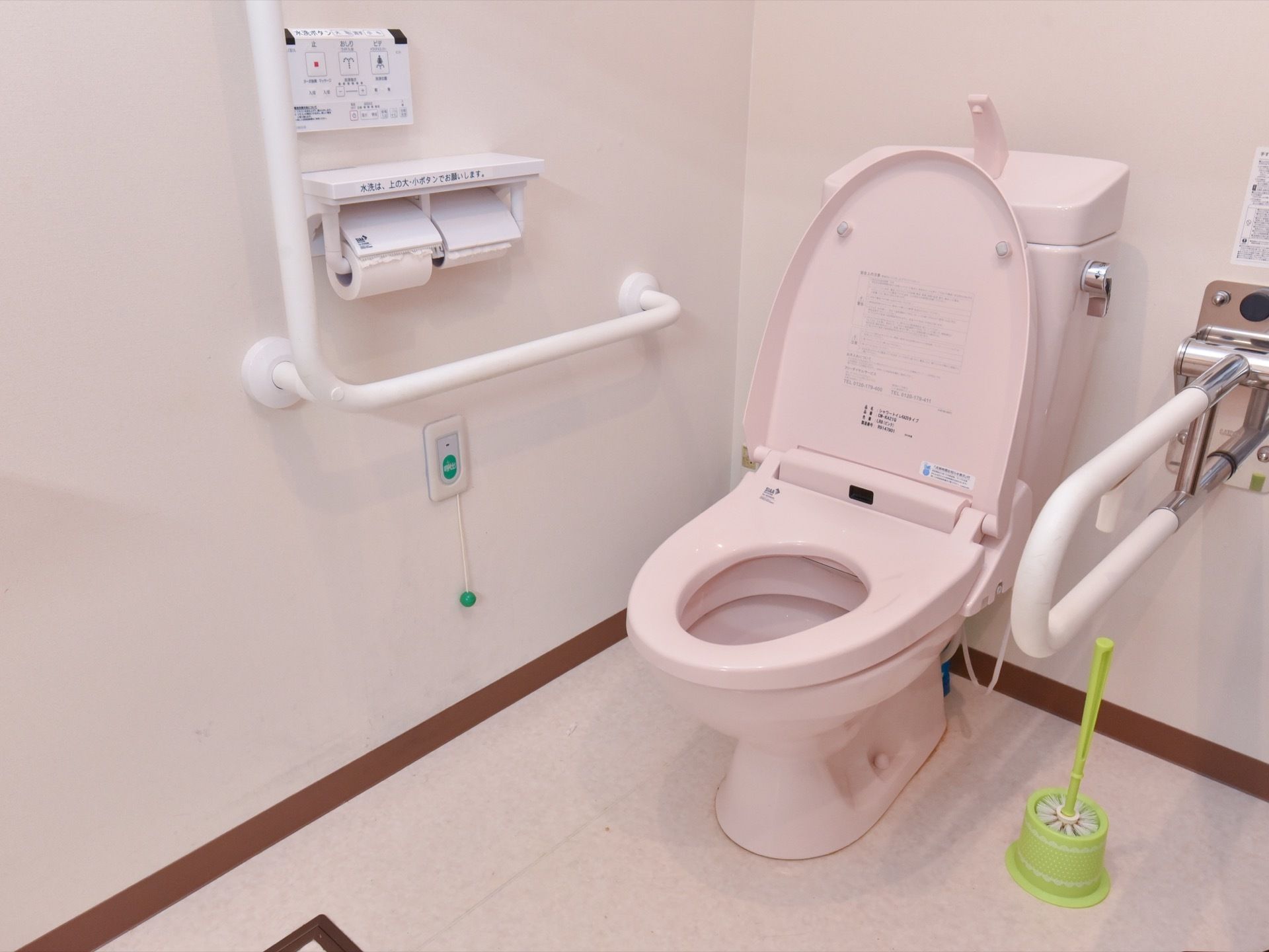 ペールピンクの優しい色合いのトイレである。左側の壁には、緊急用コールボタンがある。右下には、トイレブラシが置いてある。