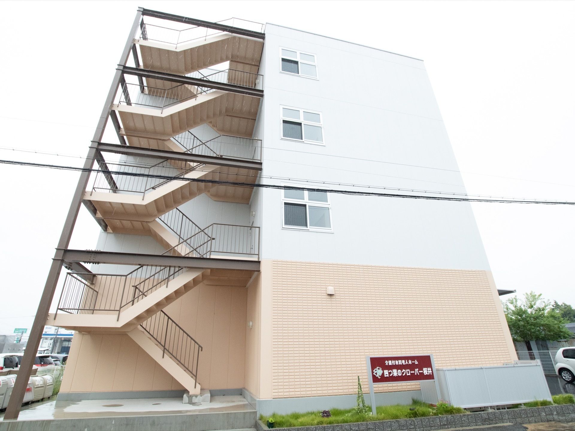 4階建ての建物が見える。正面には非常階段が見え、生垣には施設名が書かれた看板が立っている。非常階段の裏にはゴミ箱が置かれている。