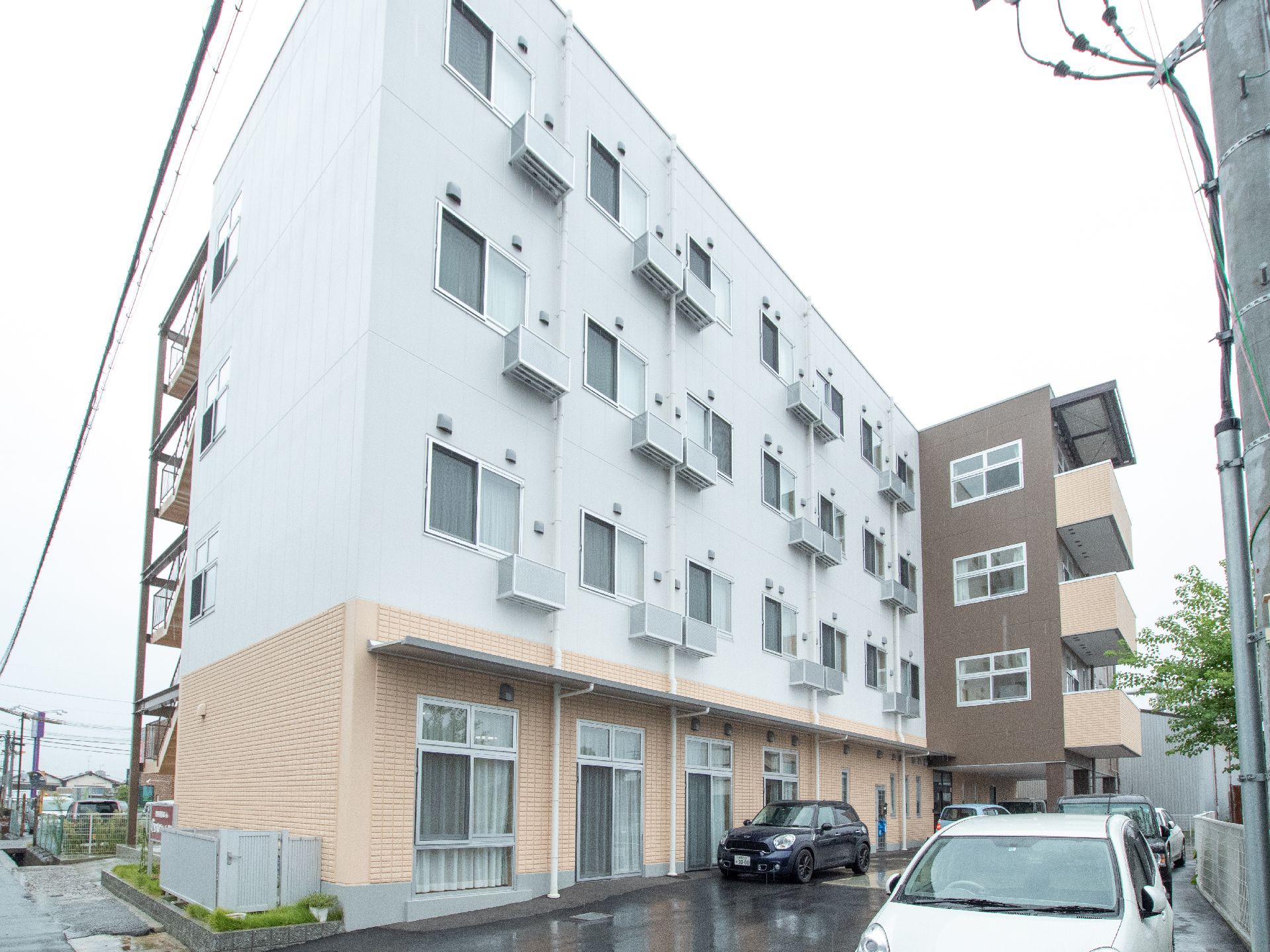 白とベージュ、ダークブラウンの外壁で、４階建てのシンプルな外観の建物である。各居室にはエアコンも完備している。