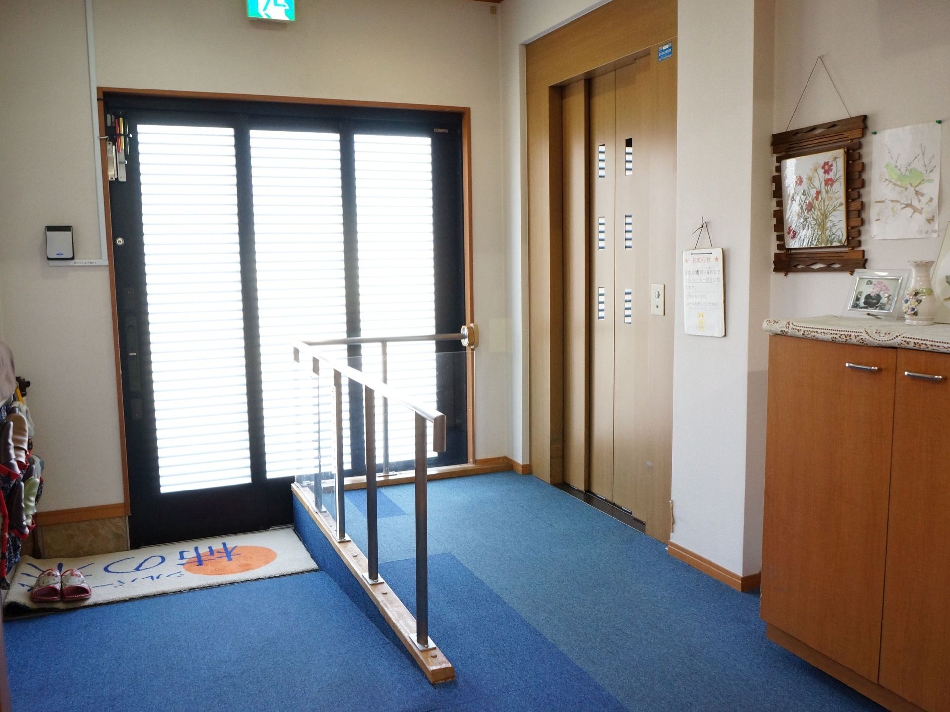 玄関を入ったところにエレベーターが見える。スロープの途中に両扉の収納棚があり、上には写真たてや花瓶が飾られている。
