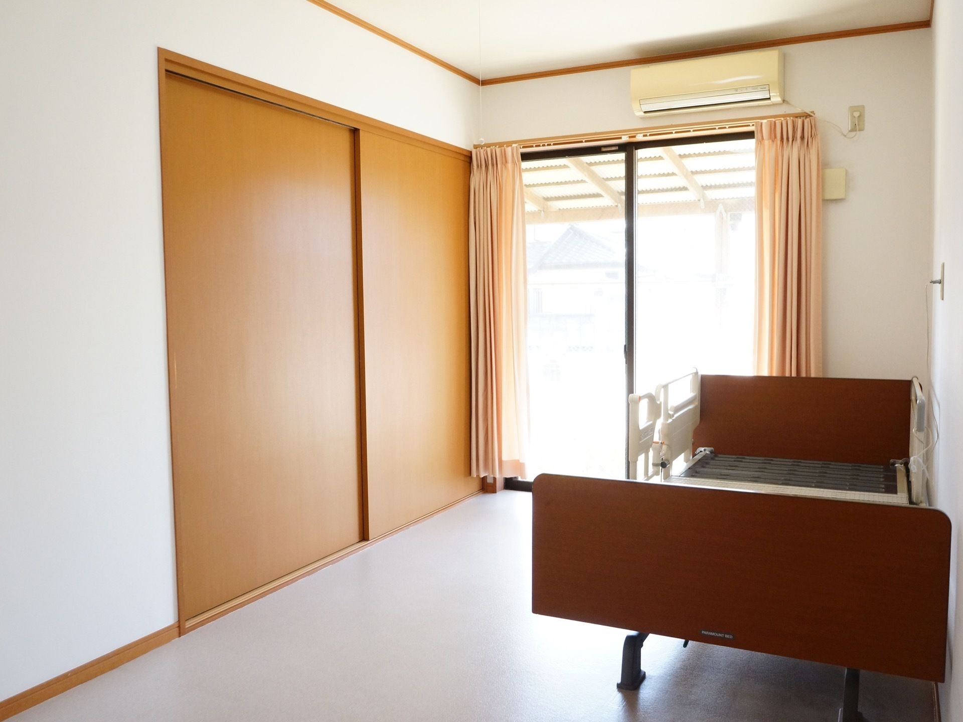 日当たりの良い、大きな収納付きの1Rの部屋。柵付きの大きな介護ベッドが置いてある。床にはカーペットが敷いてある。