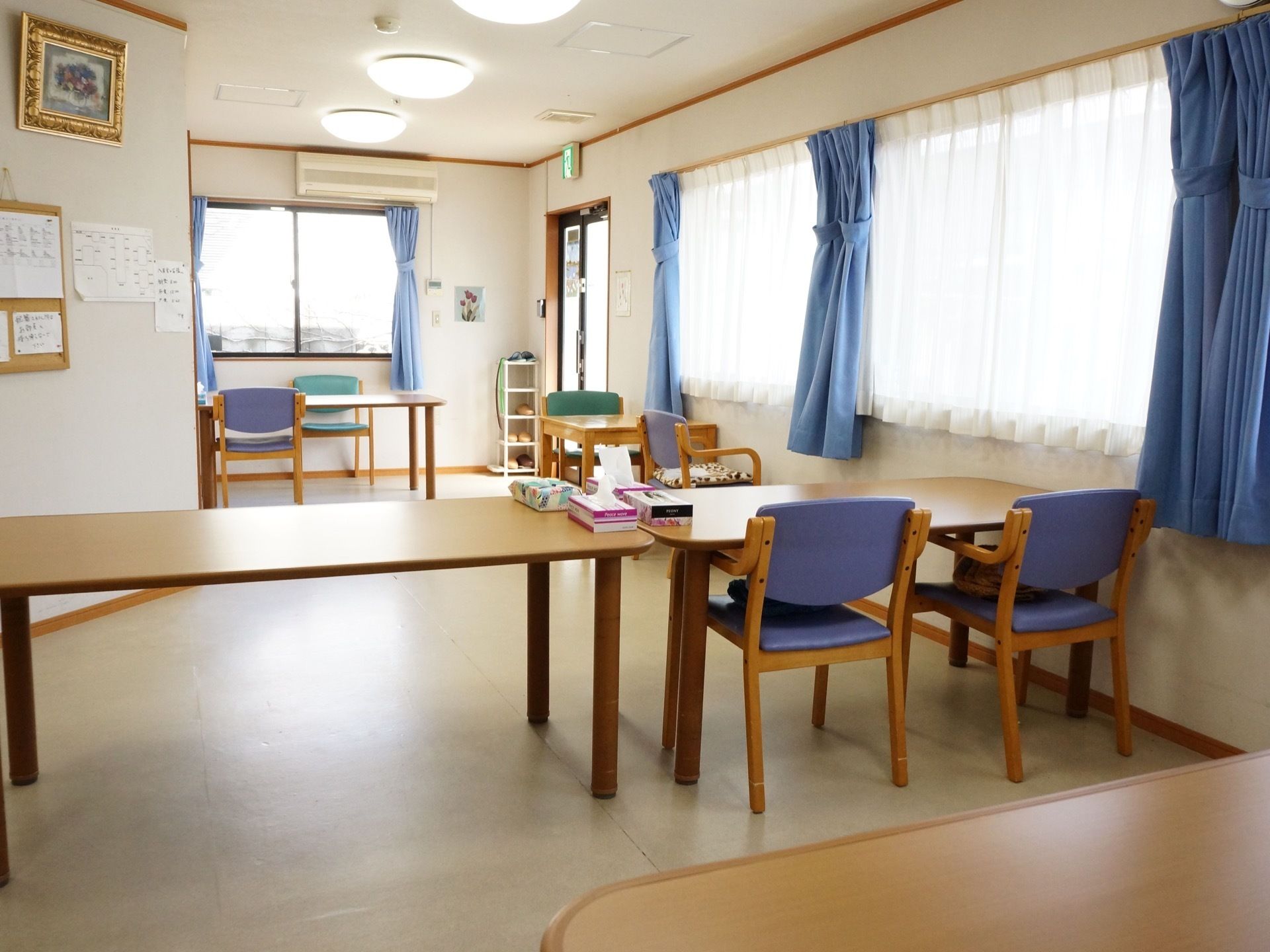ガラスの扉があり、スリッパが置かれている。窓ガラスが多数設置されていて、日差しが入っている。テーブルや椅子が置かれている。