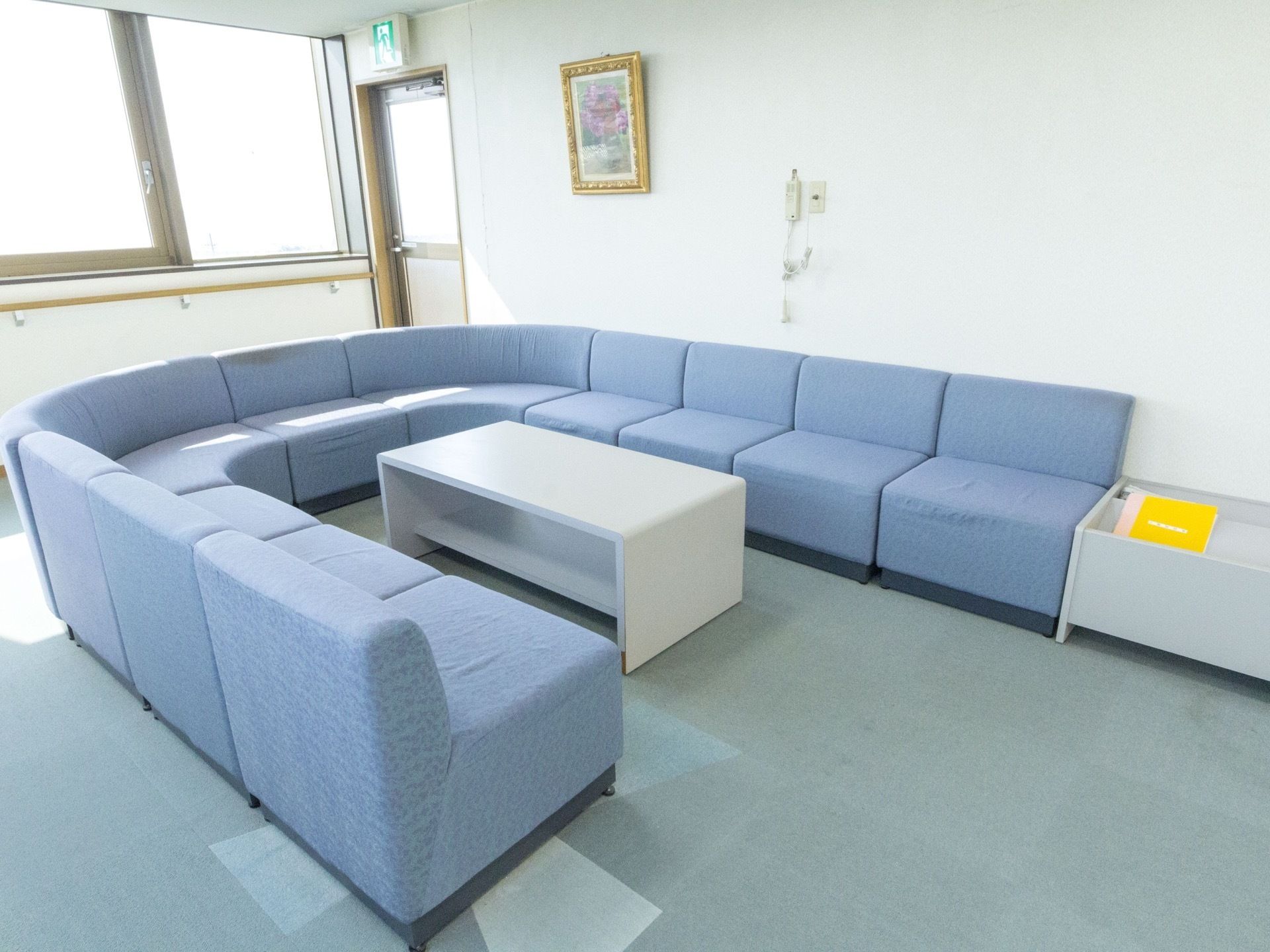 淡いブルーのソファが半円状に並べられている。中央にはテーブルが置かれている。ソファの横にマガジンラックが置かれている。