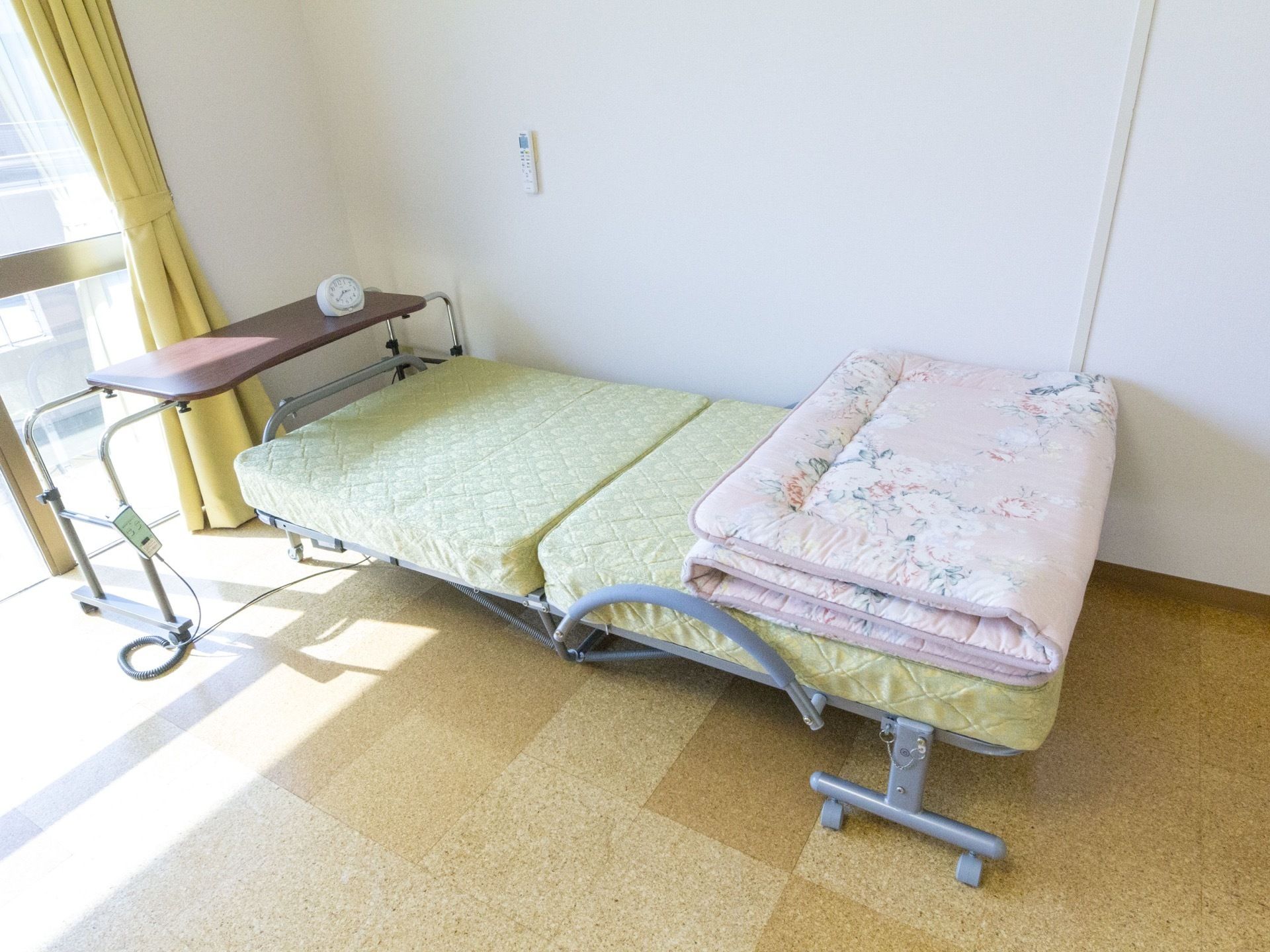 窓が見えていて、明るい日差しが入っている。ベッドが設置されていて、キャスター付きのベッドテーブルが置かれている。
