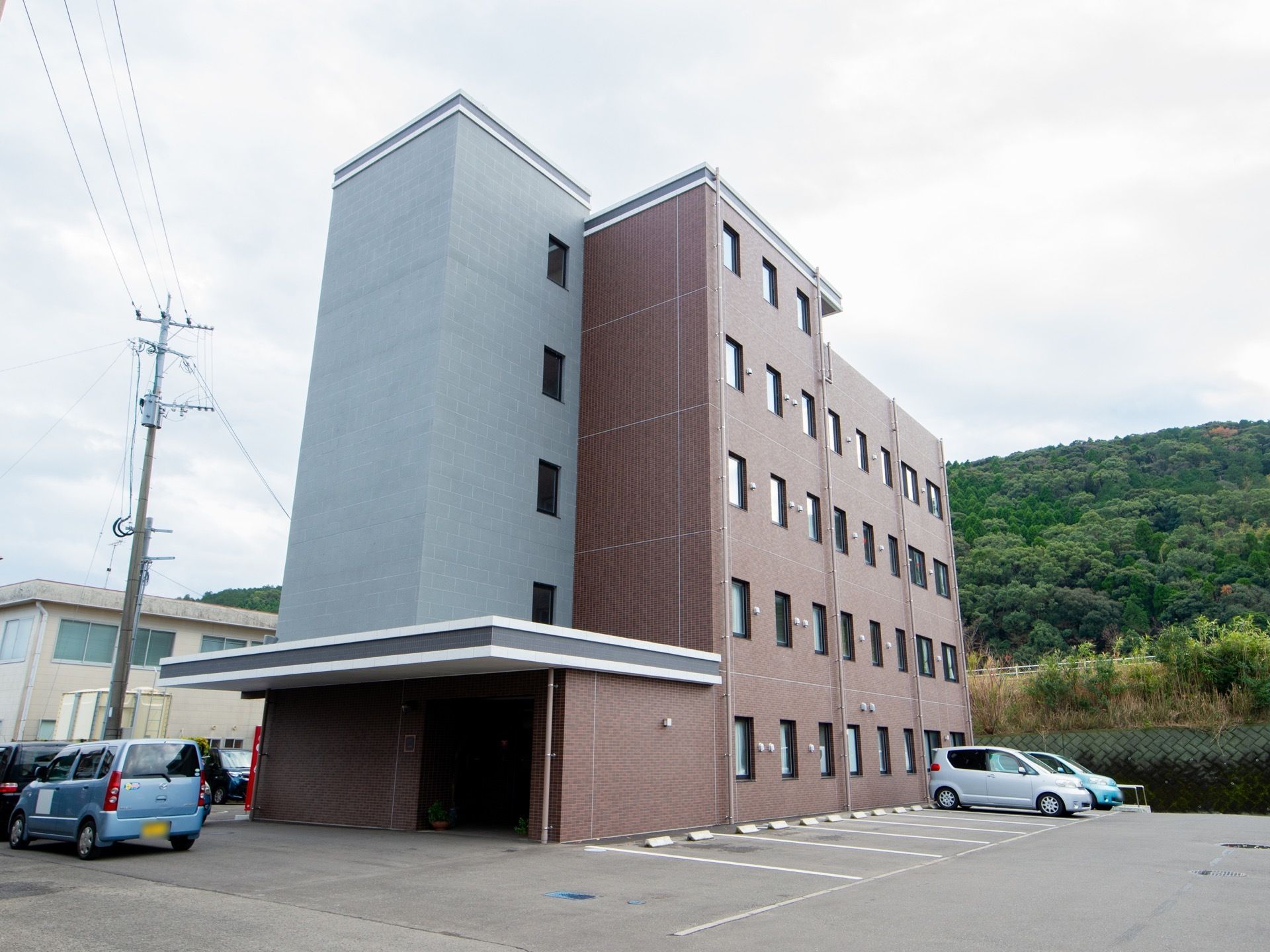 ５階建ての茶色い壁の建物で、手前に水色の壁のスペースがある。広い駐車場があり、奥側に小高い森がある。
