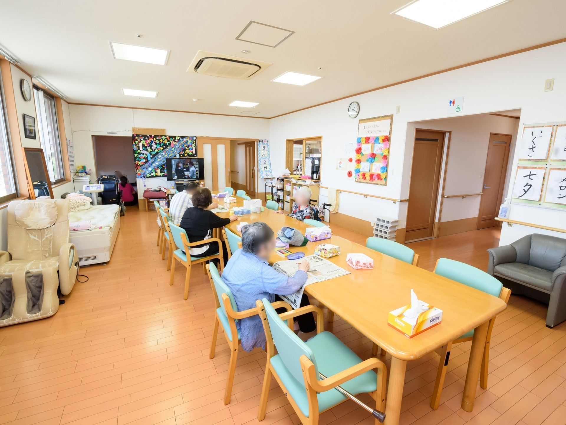 食堂にテレビやマッサージチェアが置かれている。壁には手作りの装飾や入居者様の書いた習字が貼られている。