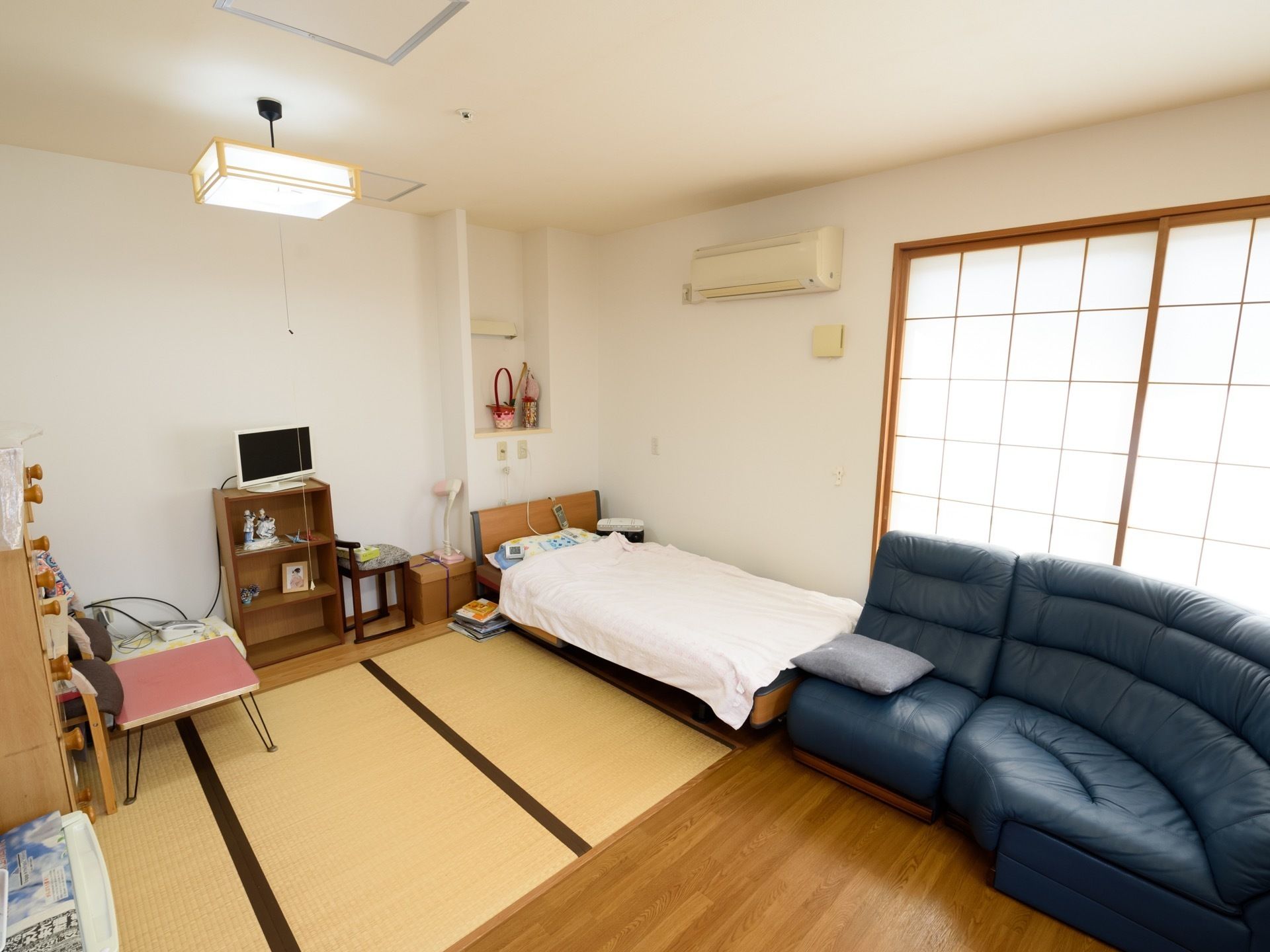 大きな障子窓があり、その手前にソファーが置かれている。ベッドは壁側に置かれており、上部にエアコンが取りつけられている。