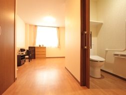 入り口を入ると、引き戸で区切られたトイレが右手にあり、奥には広い窓がある。床はフローリングで、タンスや椅子が置かれている。