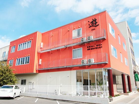 施設の建物は３階建てである。壁の色は明るいオレンジ系で、施設名が表示されている。建物の前は駐車場になっている。