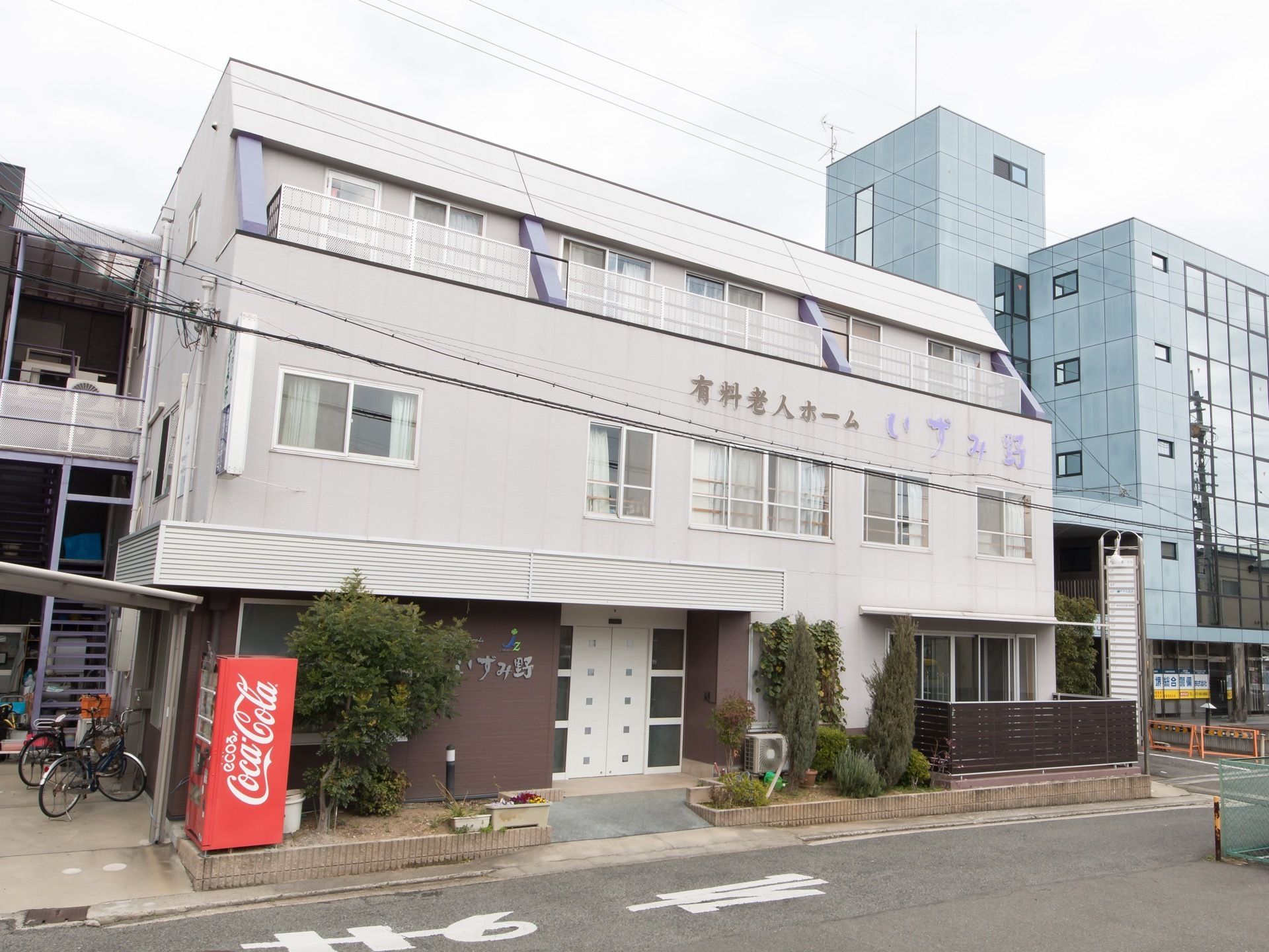全体的に白でまとめられたシンプルな外観デザインである。左端には自転車置き場があり、その横に飲料の自動販売機がある。