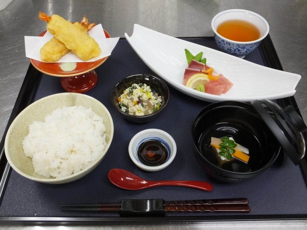 食事のメニューの一例である。膳の上には、茶碗によそわれたご飯、刺身、エビの天婦羅、小鉢、吸い物、お茶に箸が置かれている。