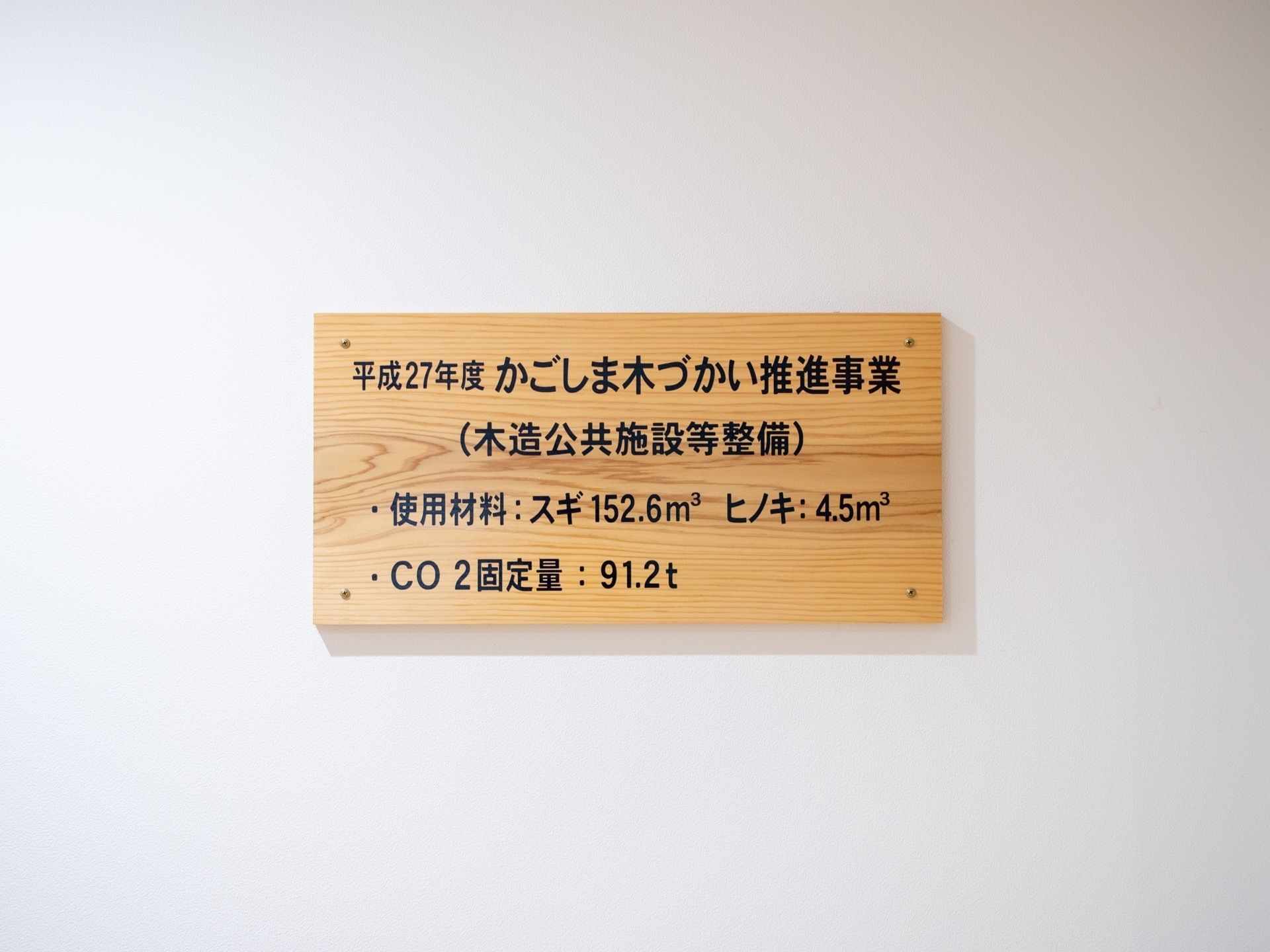 真っ白な壁に、木材の板が設置されている。施設を整備した事業名が書かれてある。また使用材料、二酸化炭素の固定量を記載。