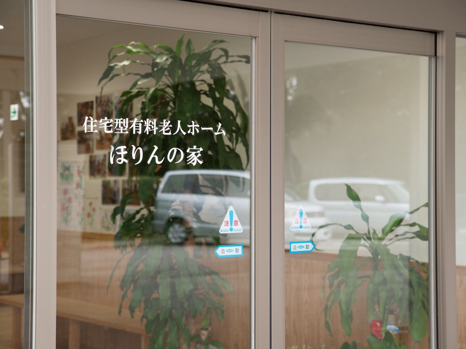 エントランスの自動ドアはガラス張りになっている。ドアには施設名が白い文字で書かれており、内部に飾られた観葉植物などが見える。