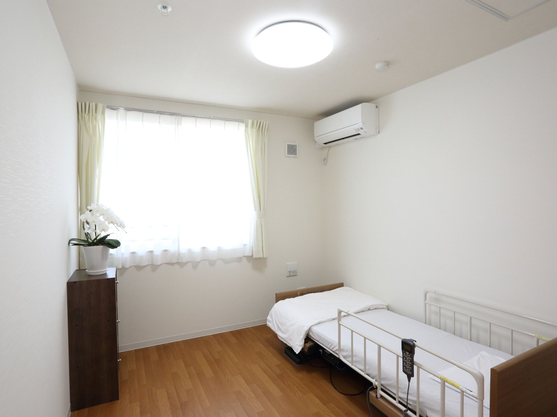 居室に介護ベッドと収納が置かれ、花が飾られている。窓が１か所あり、光が入る。エアコンが設置されている。