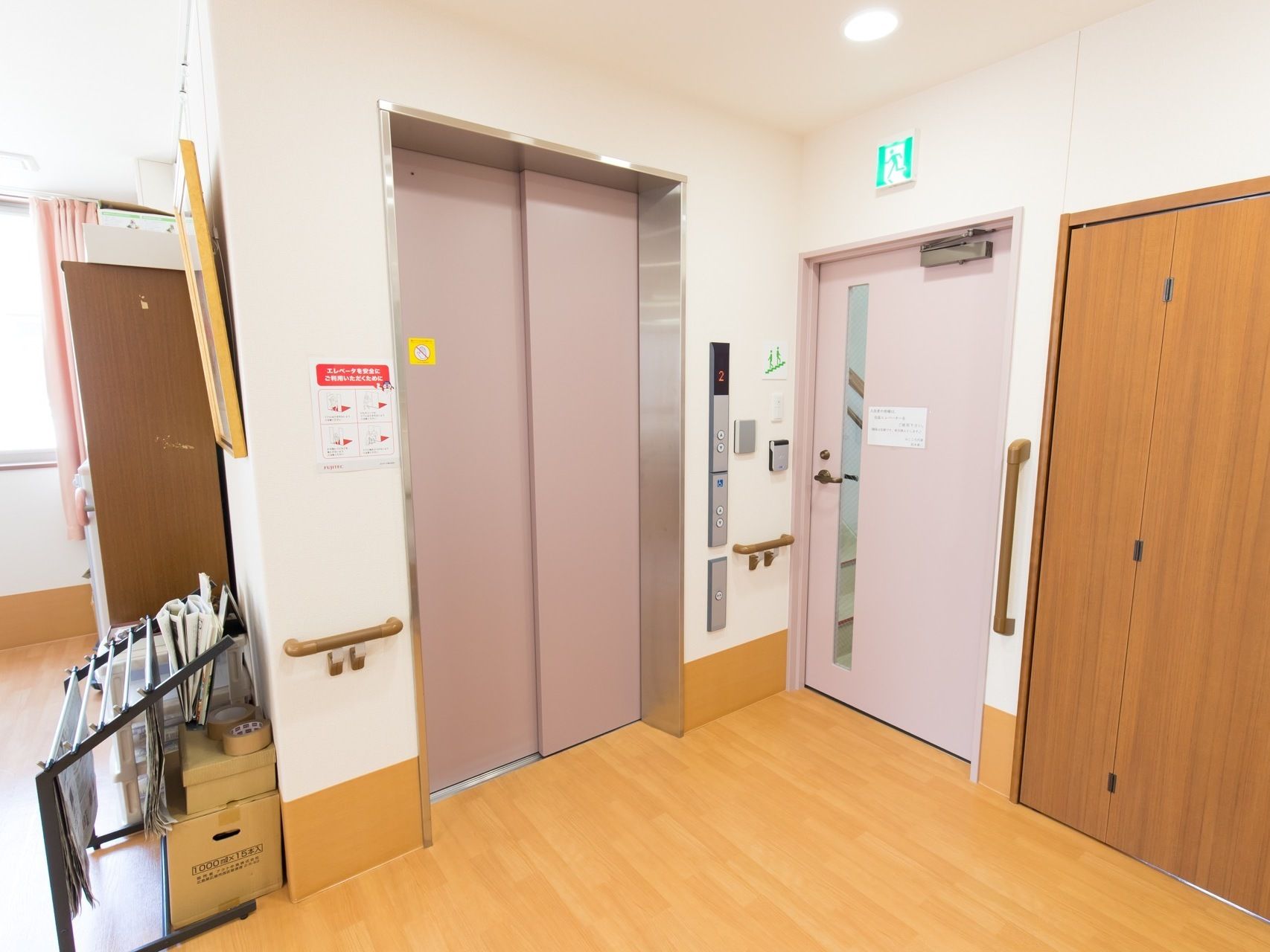 エレベーター前の通路。横には新聞ラックが置かれている。右側には非常口がある。昇降ボタンは低く設置されている。
