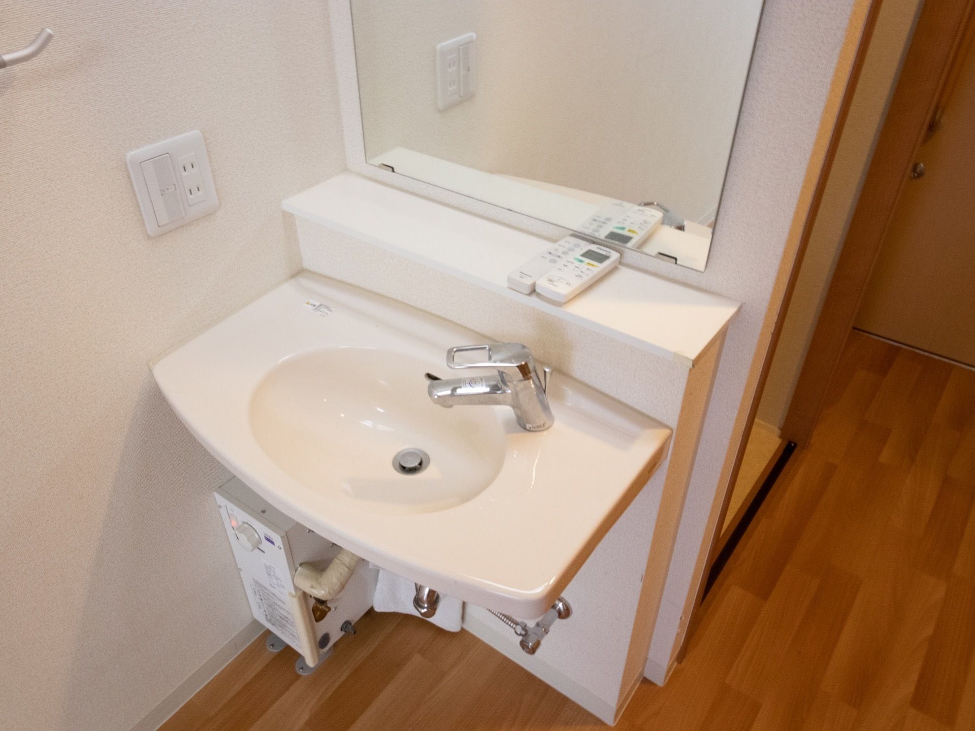 居室に備え付けられたシンプルな洗面台である。壁には鏡が付いており、前の棚にはリモコンが置かれている。