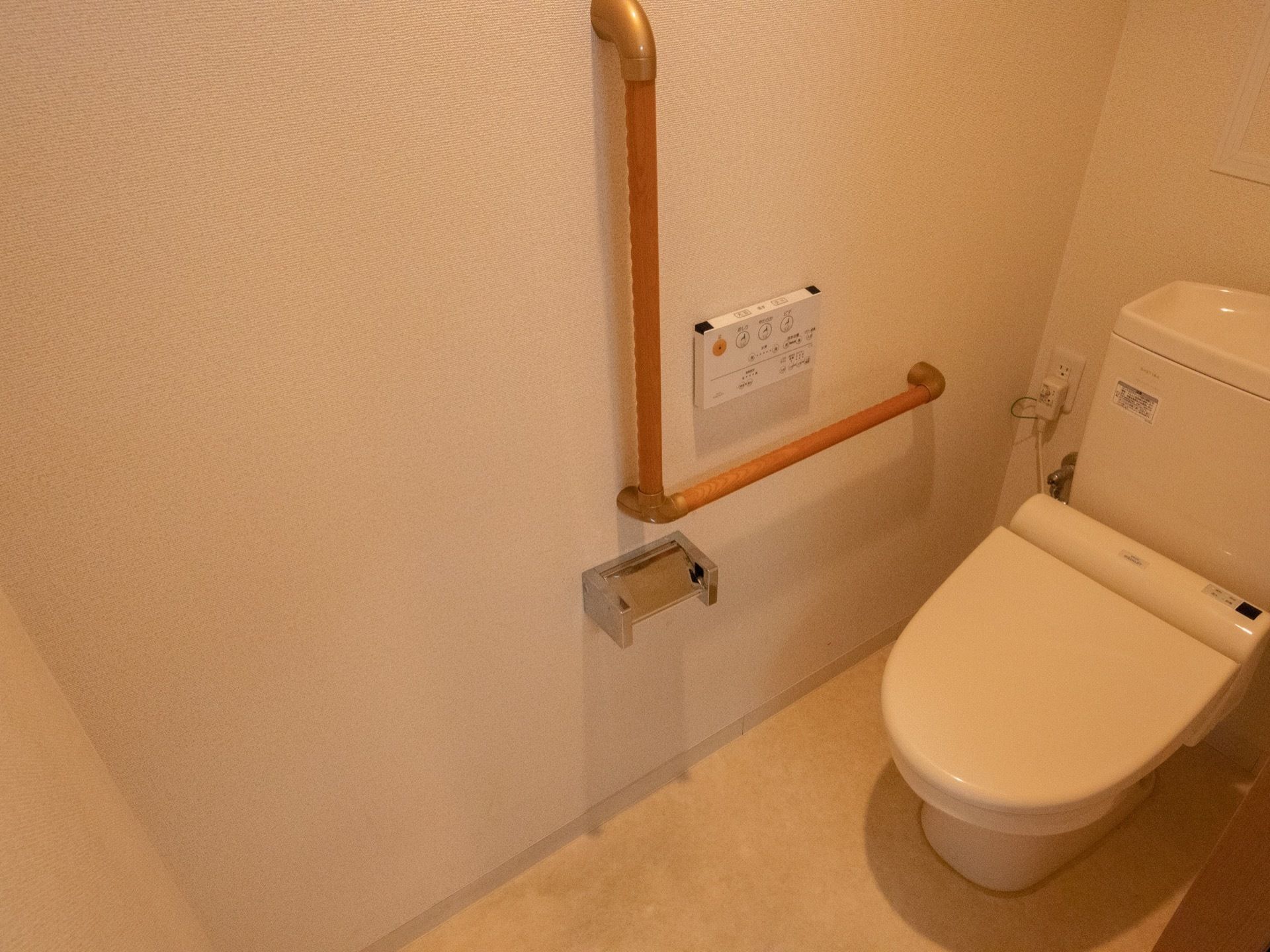 温水洗浄機能付きの洋式トイレである。横の壁には操作パネルやＬ字型の手すり、ペーパーホルダーが取り付けられている。
