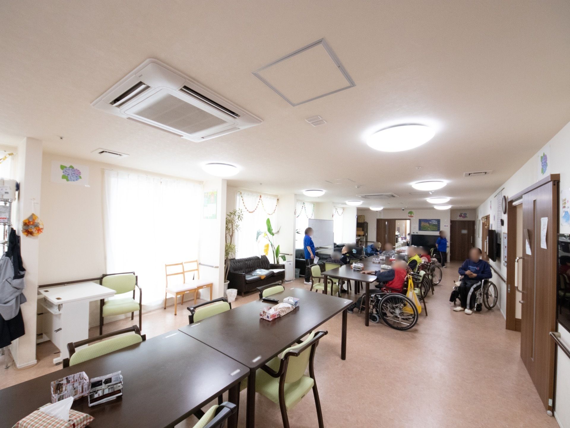 部屋の中央にダイニングテーブルと椅子が数脚並べて置いてある。数名の車椅子をご利用の方が青い服のスタッフの話を聞いている。