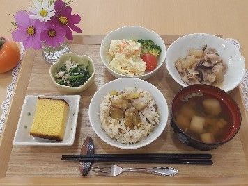 木製のプレートに季節ご飯と一汁三菜の献立が並べられている。栄養バランスが考慮され、食べ応えのあるメニューである。左上には花が飾られている。