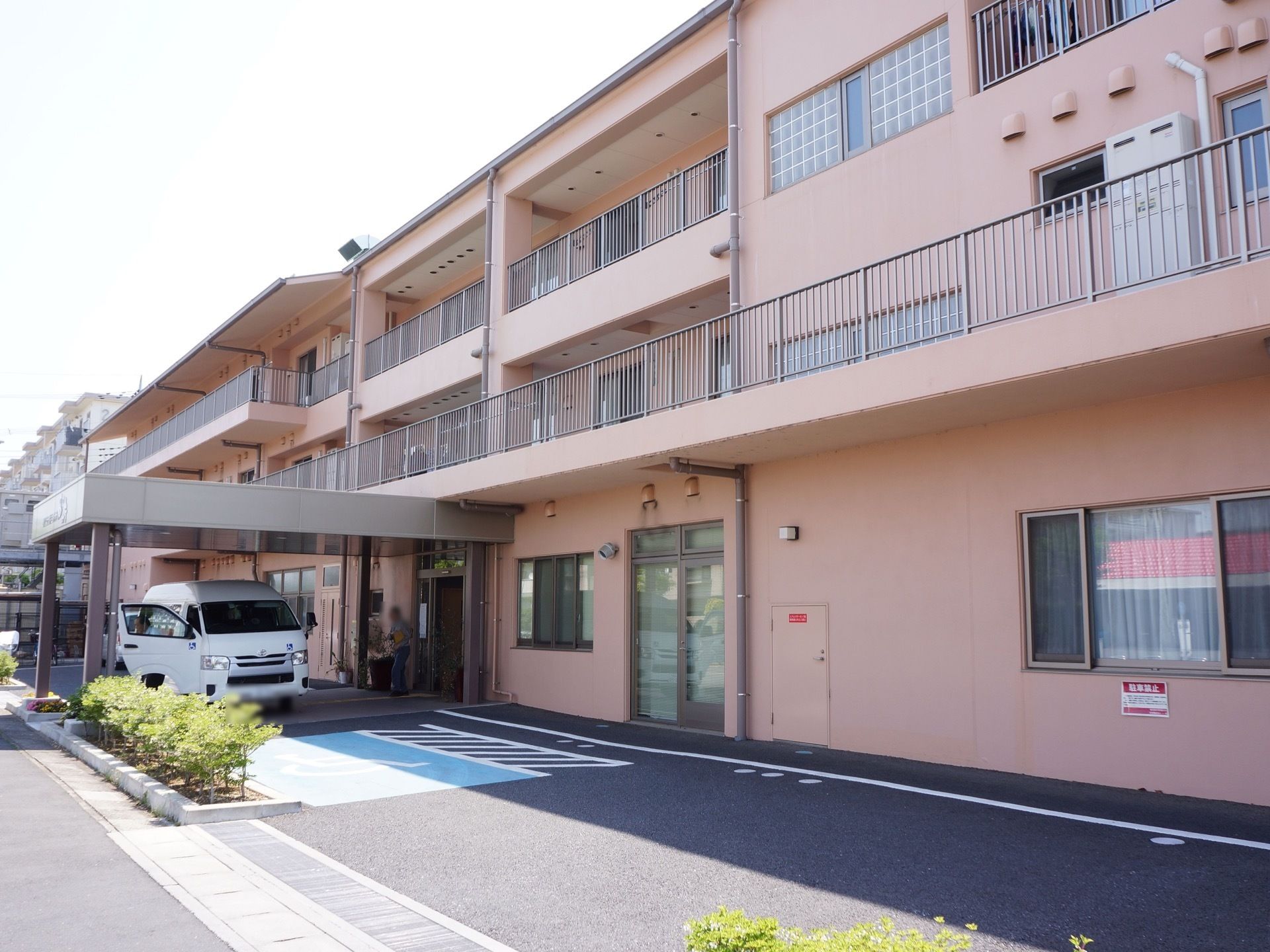 ベランダのある淡い桃色の建物である。路面はアスファルトで舗装されていて、入り口前にはワンボックスの車が停車している。