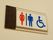 壁に取り付けられたトイレの場所を示すプレートの写真。木製のプレートに文字ではなくマークで表示されている。