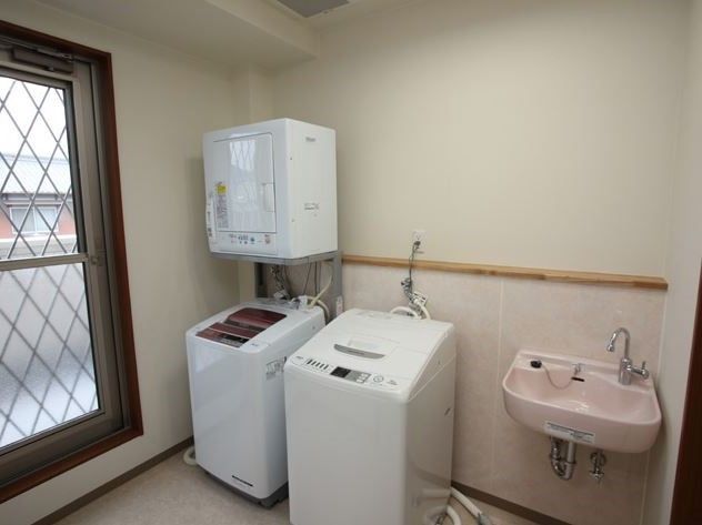 ２台の洗濯機と、１台の乾燥機が設置されており、横には水道がある。清潔感のある白い壁で、窓が大きく明るい。