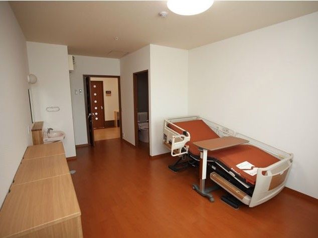 介護用ベッドが置かれた室内の様子。開いているドアの奥にはトイレが見えている。木目調の背の低い家具が設置されている。