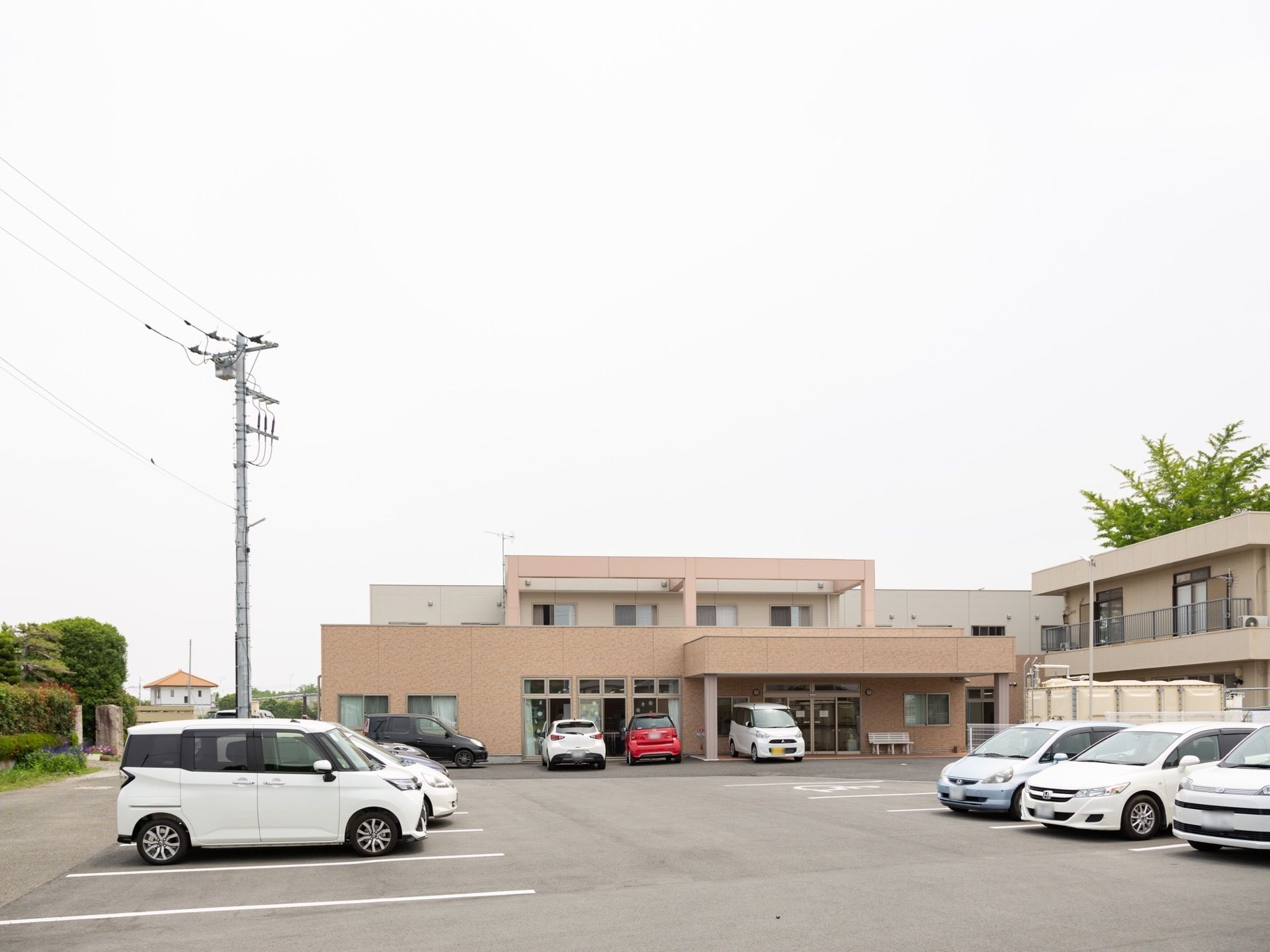 施設は茶色の壁をベースとした２階建てのコンパクトな建物である。施設正面には駐車用のスペースがあり、数台の車が停まっている。