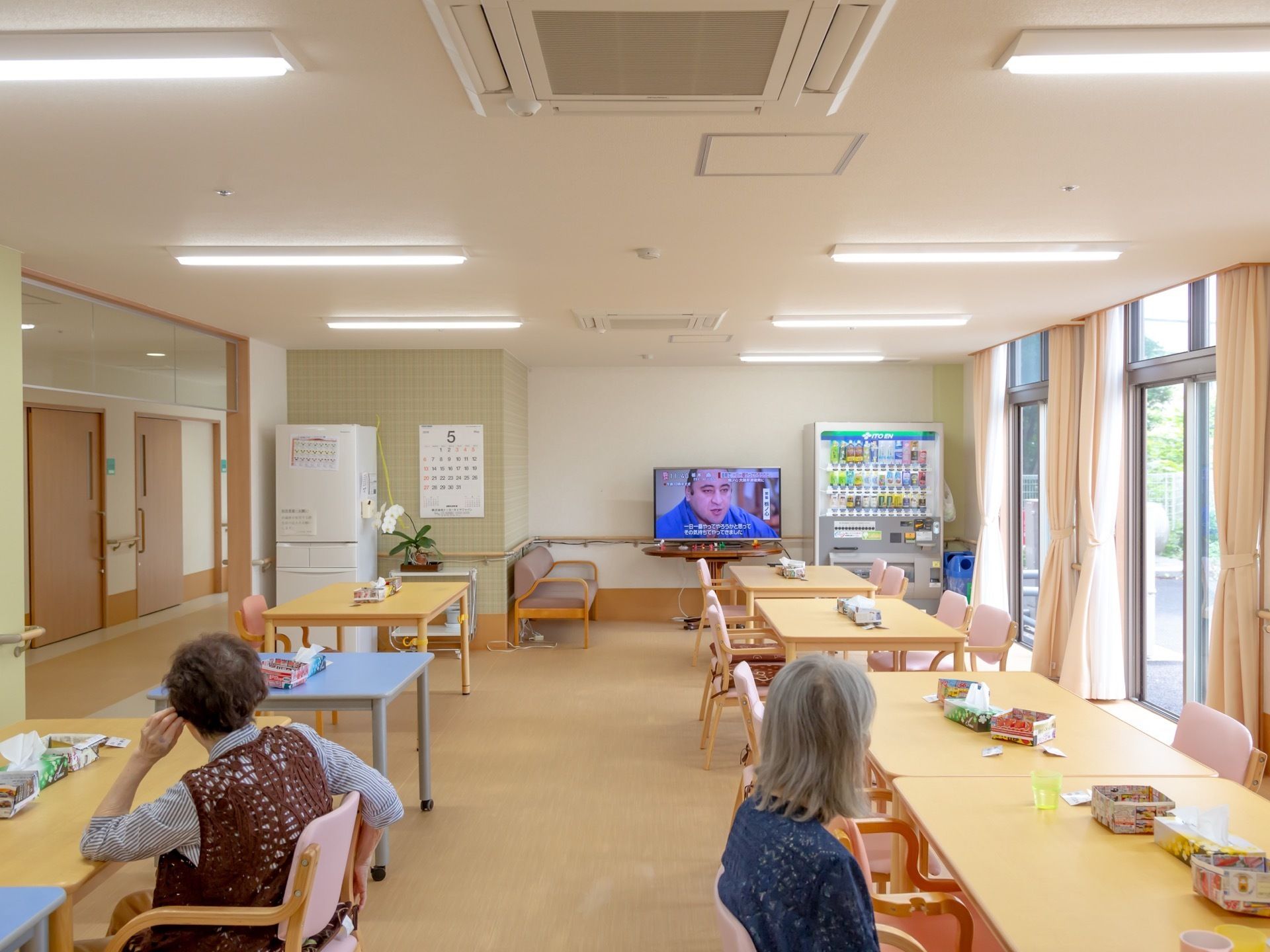 大きめの長方形のテーブルが二列で整然と並べられており、周囲には椅子が揃っている。入居者様が椅子に腰を掛けてテレビ観賞している。