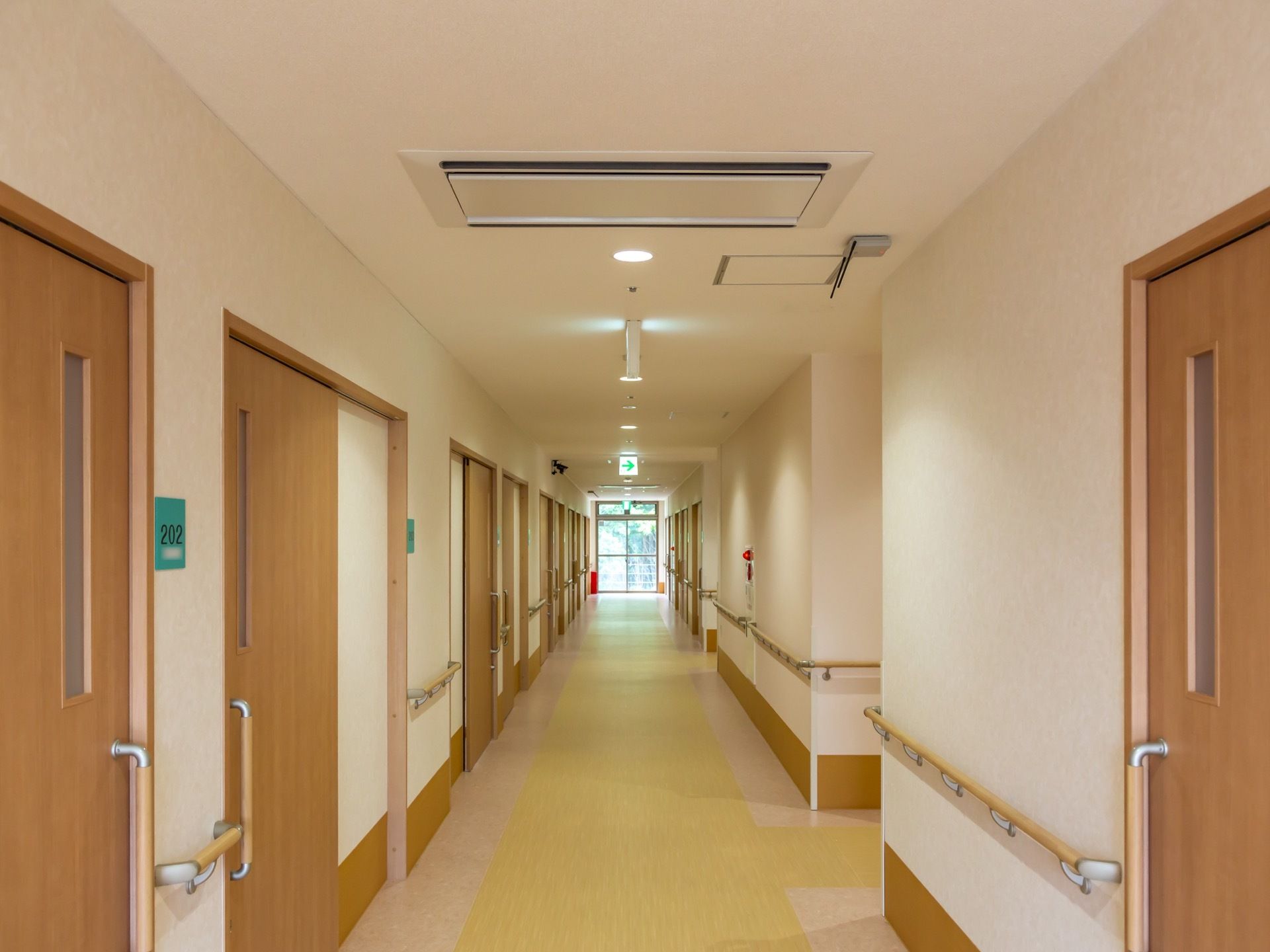 明るめの茶色やベージュであしらわれた、入居者様の居室が一続きに並ぶ廊下である。両側の壁には手すりが備え付けられている。