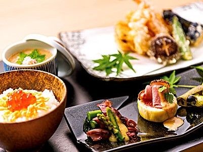 黒いお盆の上に、天ぷらの盛り合わせや茶碗蒸しなどの和食が並んでいる。葉っぱをちらして季節感を演出している。