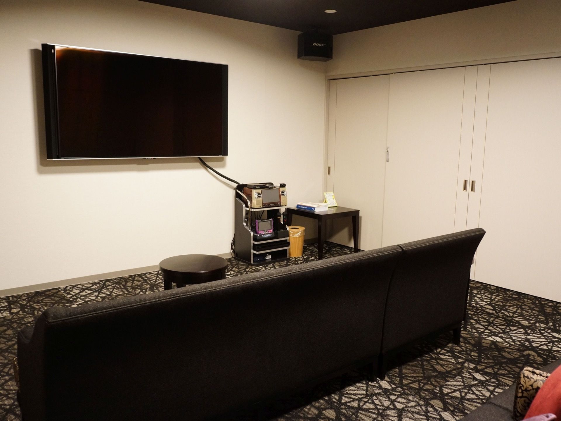 部屋の中央に黒いソファーが置かれ、壁には薄型テレビが掛けられている。カラオケセットとゴミ箱とコーナーテーブルがある。