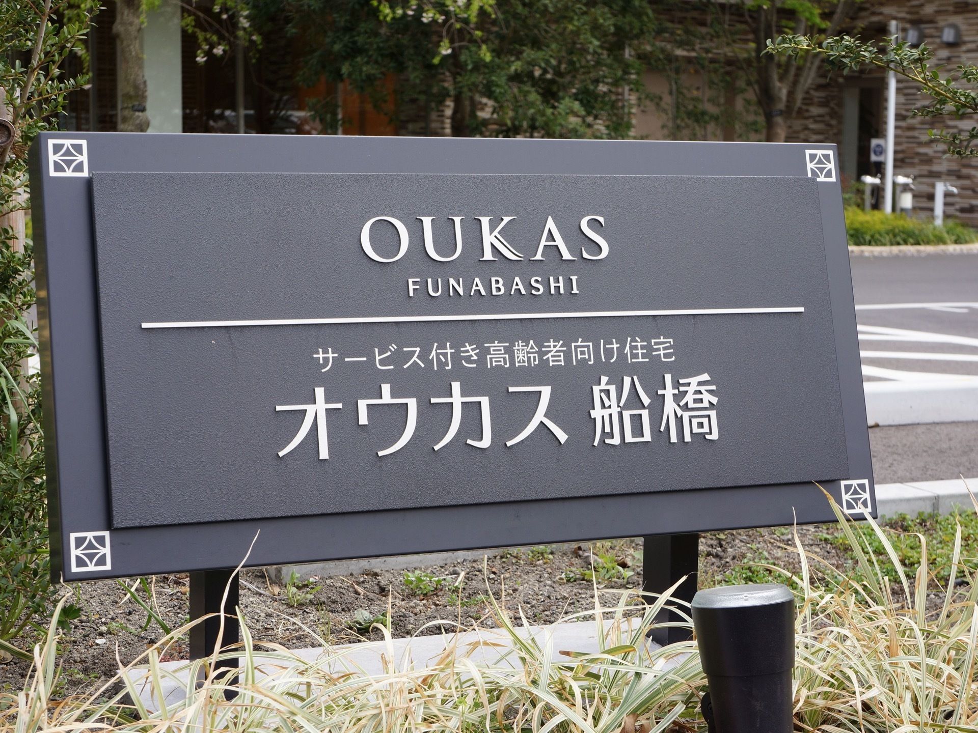 黒い看板に銀色の文字で施設名が表示されている。看板を照らすライトアップ用の照明が地面に設置されている。