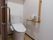 フローリングの床の上にタンク付きの白い洋式トイレが設置されている。便器の上に棚が取り付けられ、トイレットペーパーなどが置いてある。