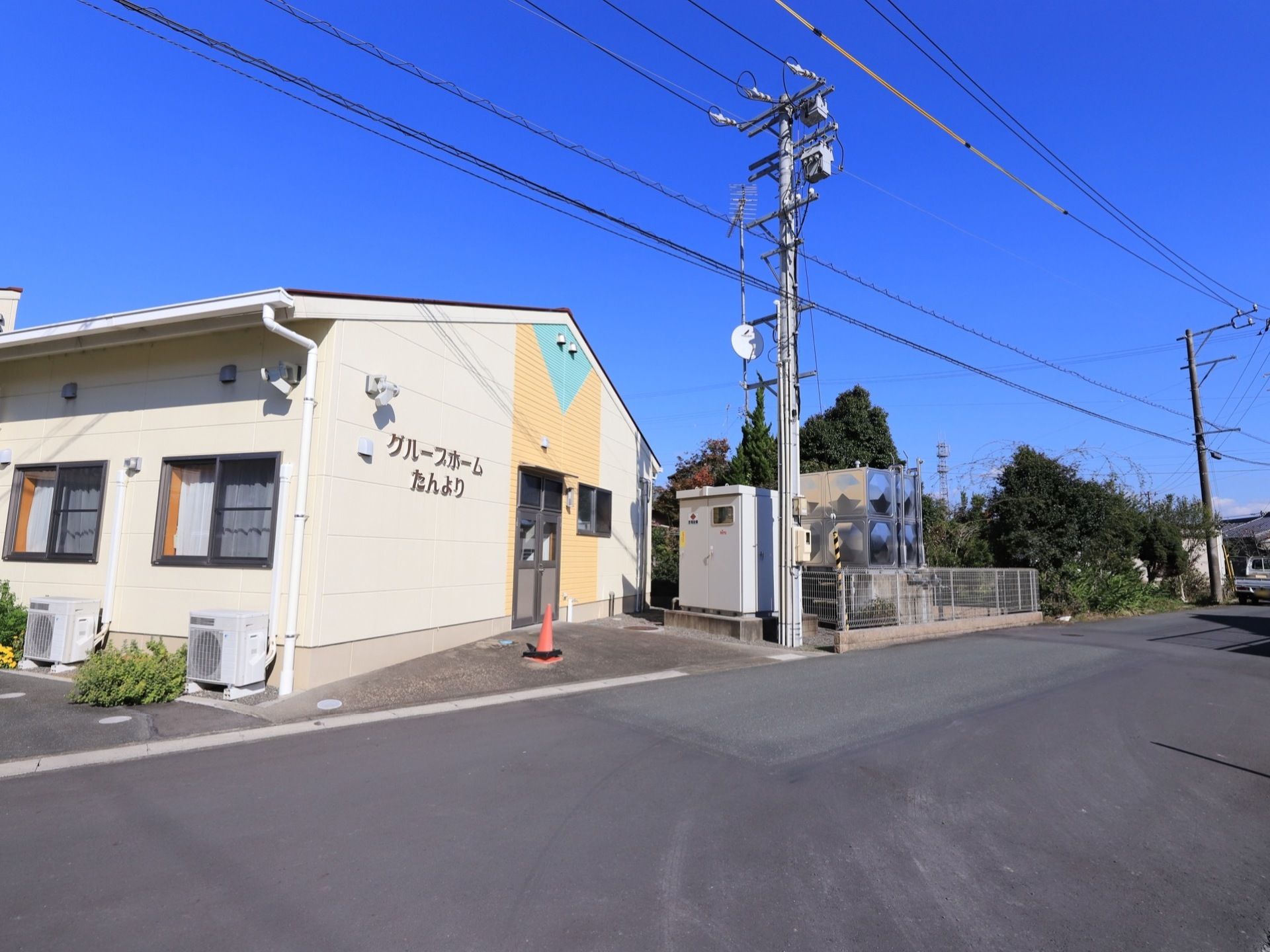 施設は白色の外壁で、居室の外にはエアコンの室外機が置かれている。施設名書かれた壁のそばには、扉が設置されている。
