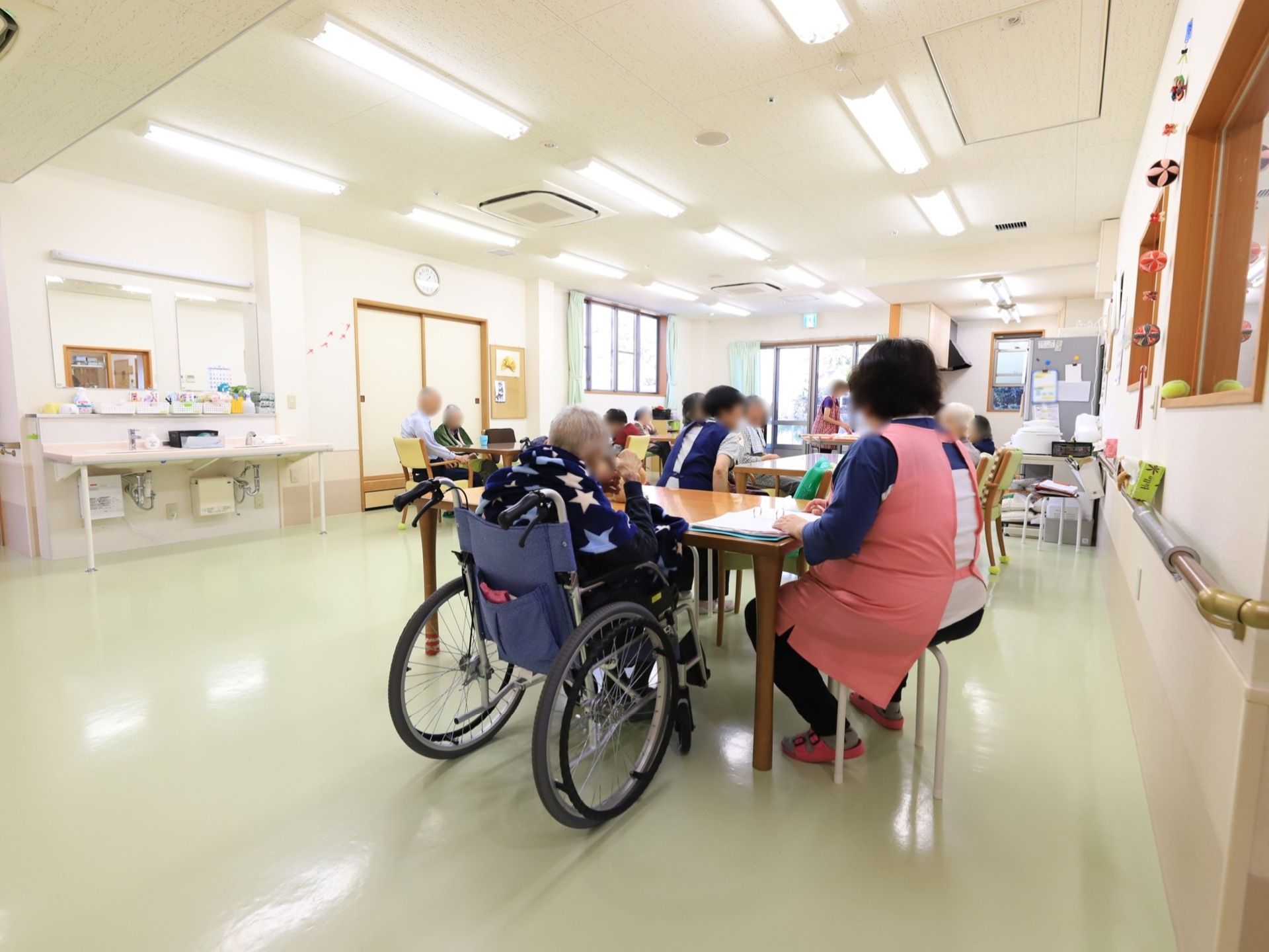 共有スペースは広々としており、大勢の入居者様とスタッフが一緒に生活をしている。天井には、大きなエアコンが2ヶ所設置されている。