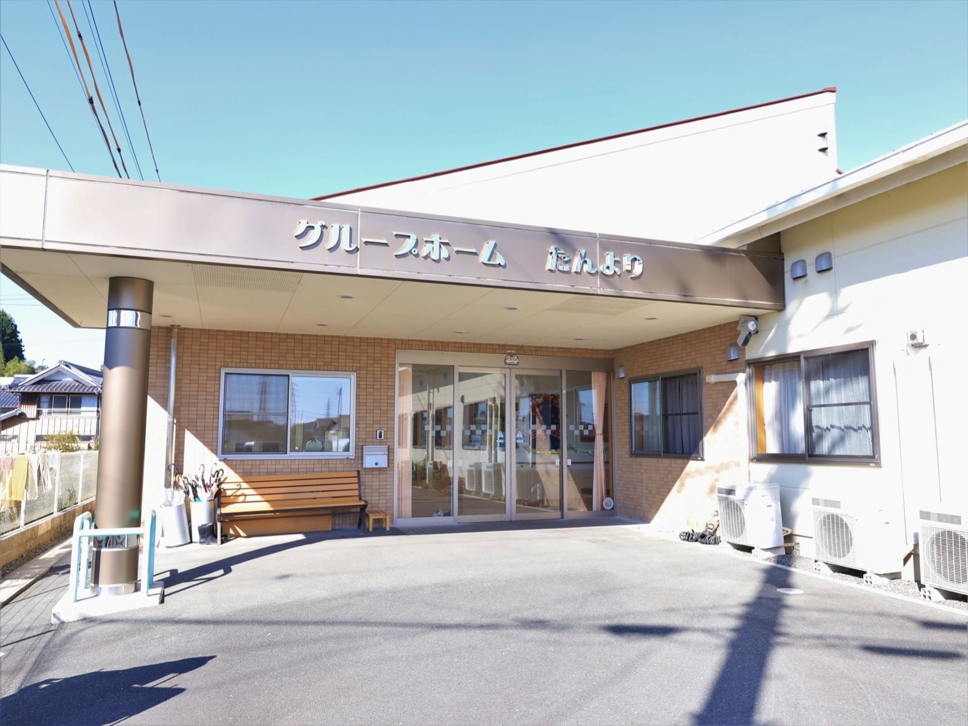 施設の玄関前には、茶色くて大きな屋根が設置されている。その下には、ベンチや傘立てなどが置かれている。