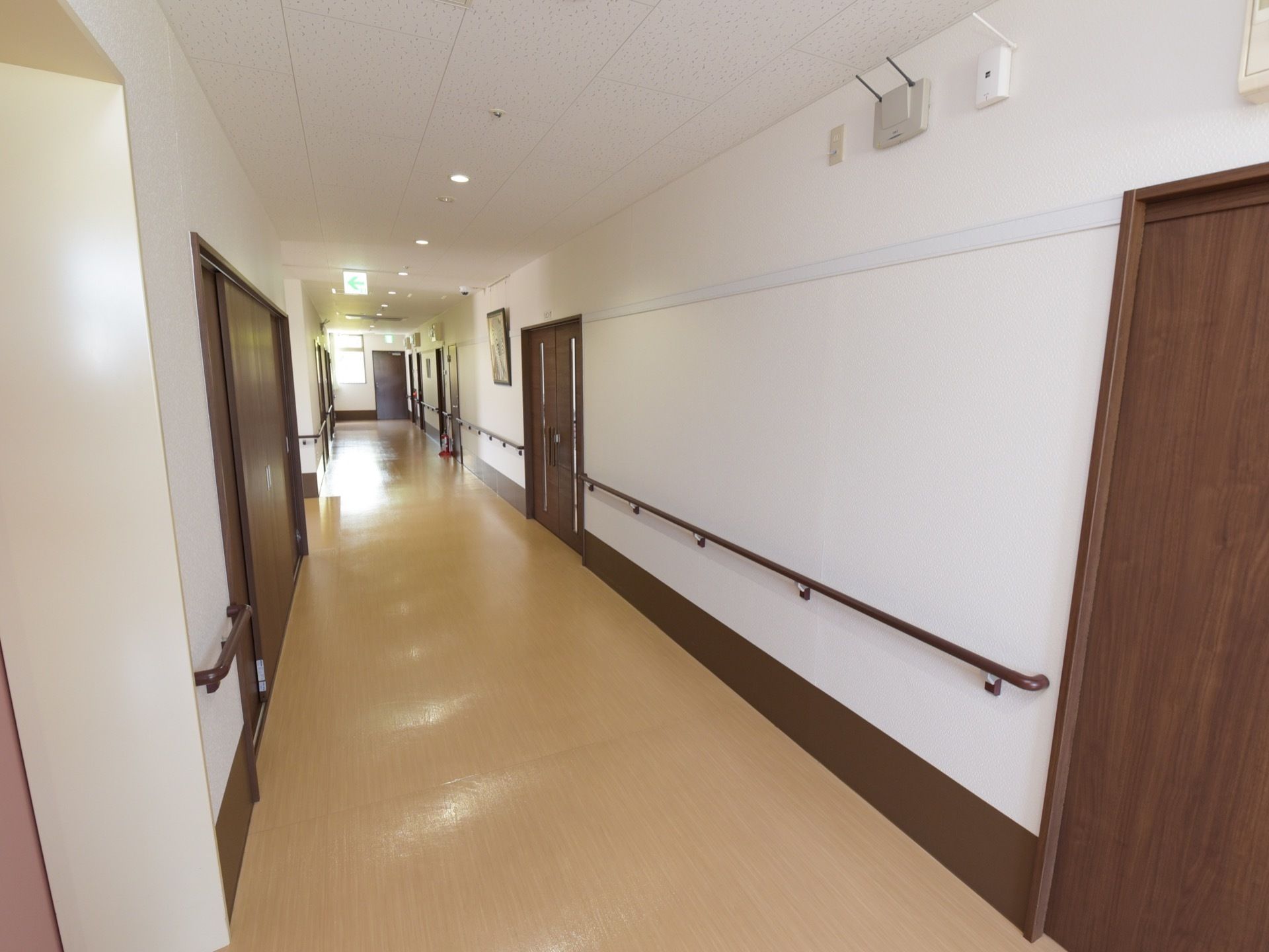 壁には手すりが設置してある。非常口灯と消火器がある。壁は白色でドアは焦げ茶色の配色である。床はバリアフリーである。