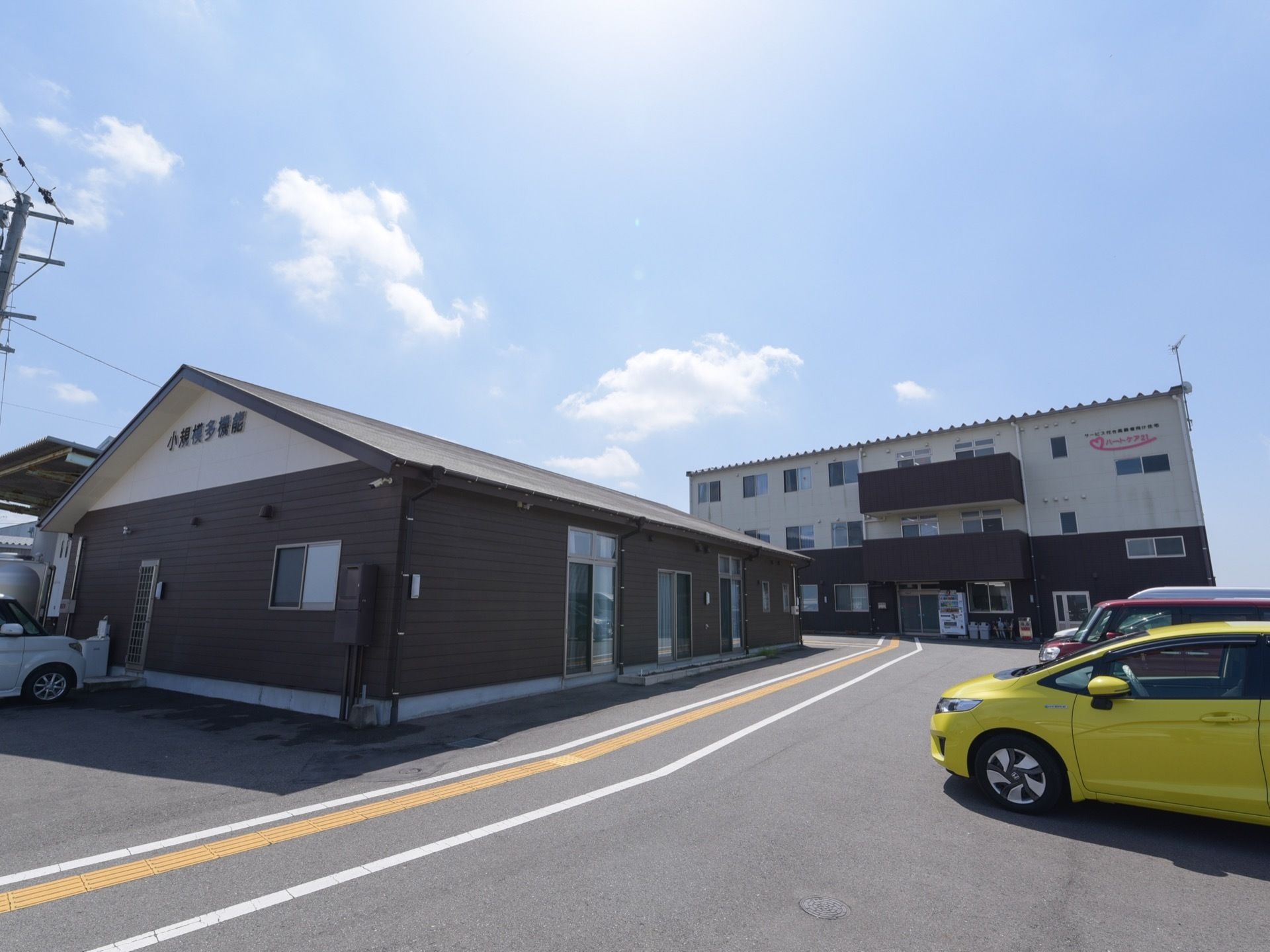 アイボリーとこげ茶色の配色の外壁である。手前に別施設がある。奥に3階建ての施設がある。駐車場が完備されている。