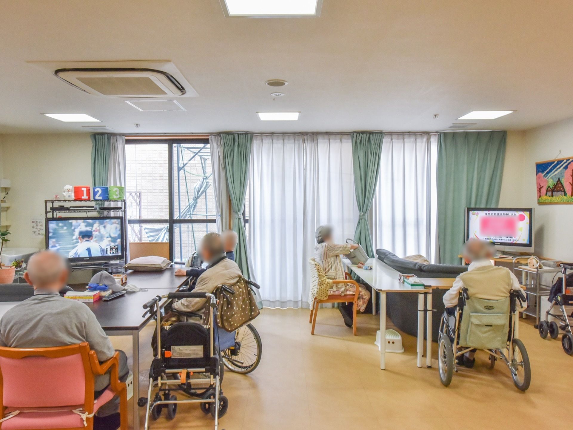 共有スペースは壁一面に大きく窓が設けられている。テレビが２台置かれ、うち１台のテレビ前はＬ字型のソファが置かれている。
