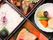 重箱に詰められた食事で時計回りにおかず、煮物、焼き魚、ごはんが入っている。ごはんには小さな漬物が添えられている。
