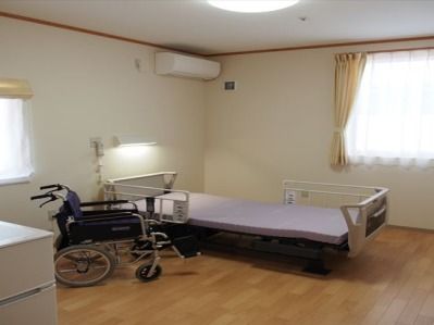 居室はフローリングの床となっており、中央に配置された柵のついたシングルベッドのそばに車椅子が置かれている。